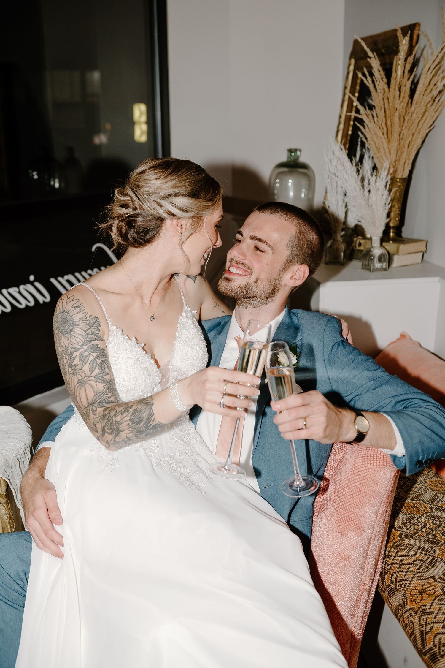 A couple in wedding attire share champagne while sitting together on a pink chair with decorative pampas grass nearby.