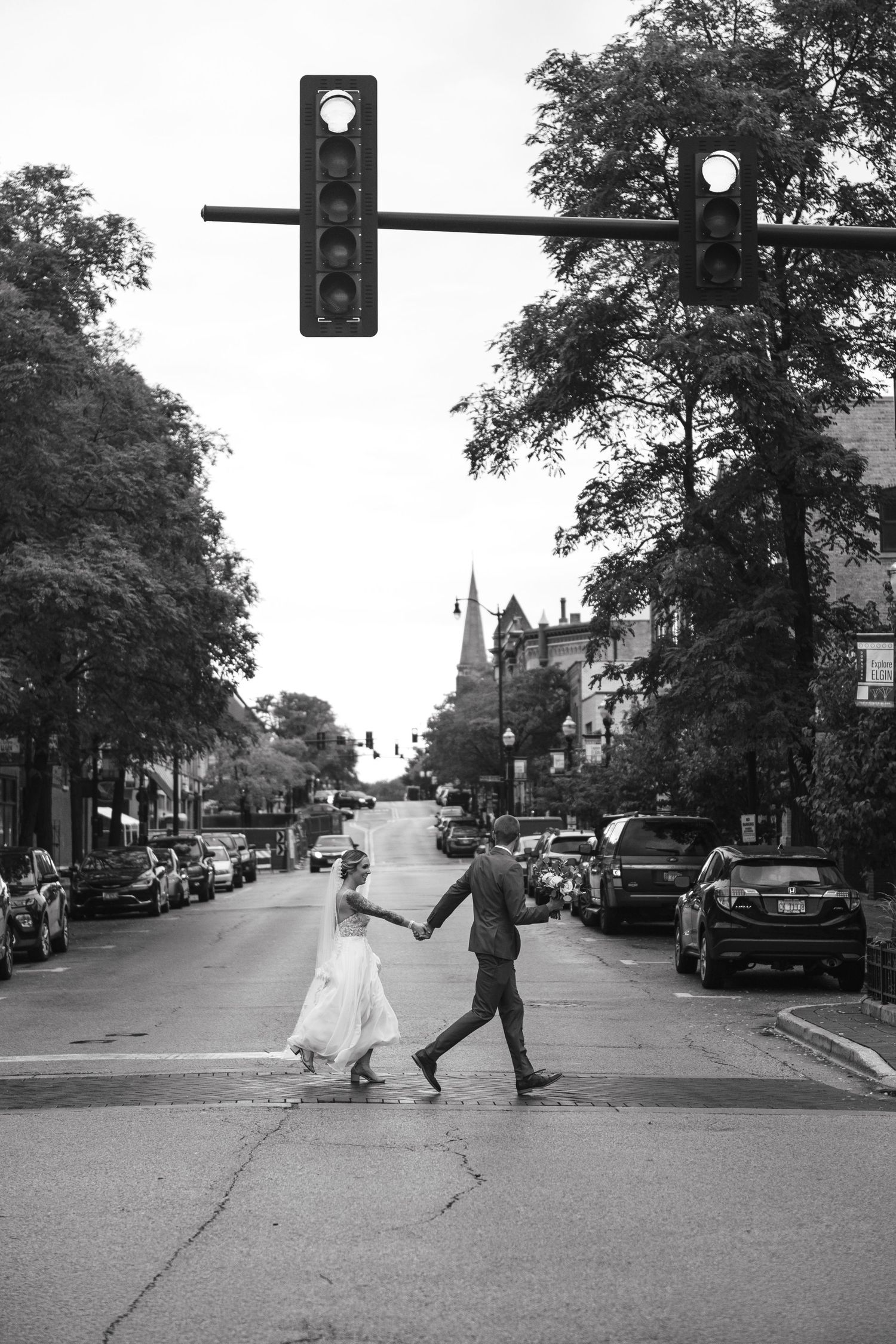 A couple crosses a tree-lined street in a charming downtown area with parked cars and traffic lights above.