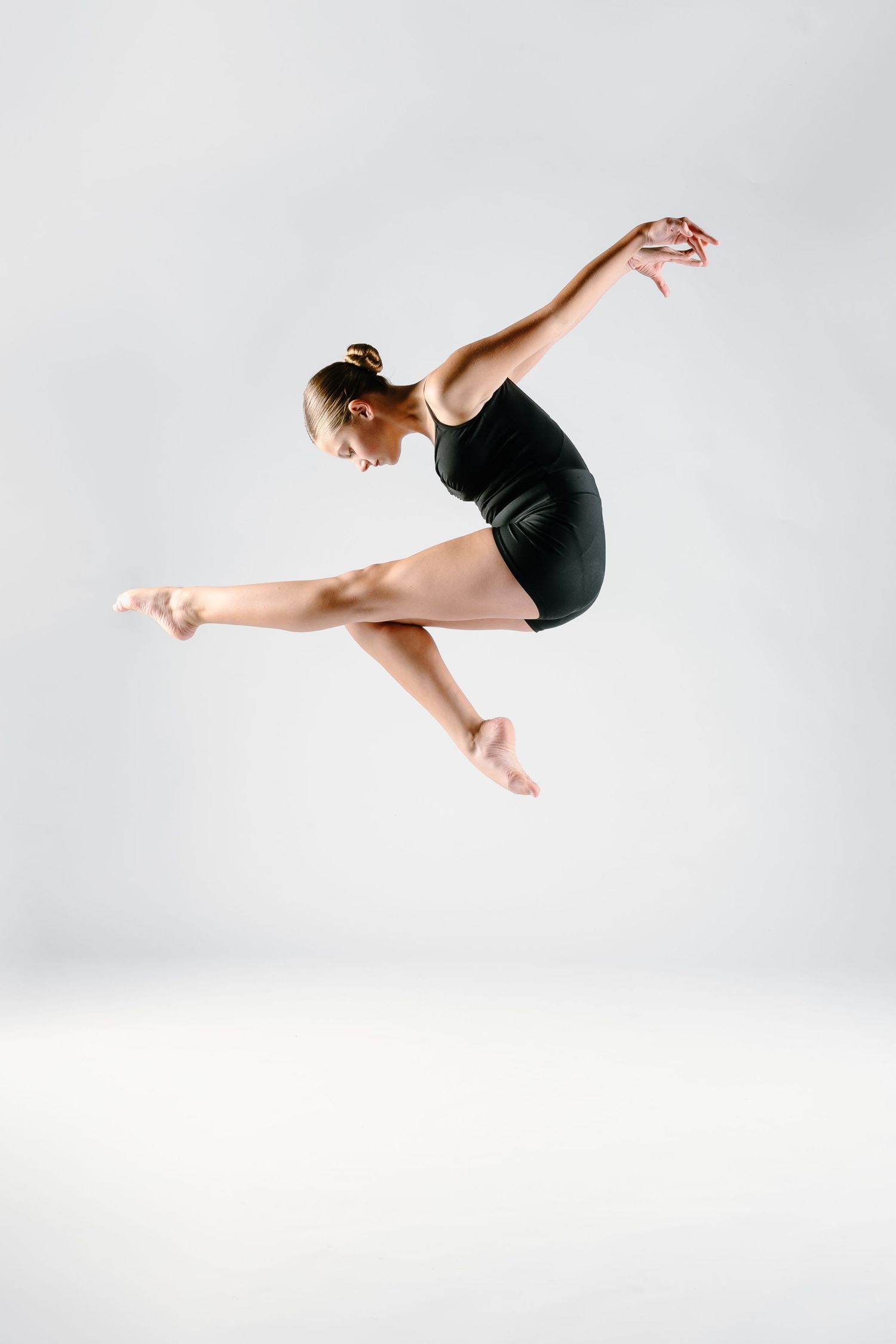 Female dancer in black leotard performing a graceful jump with outstretched limbs against white background.