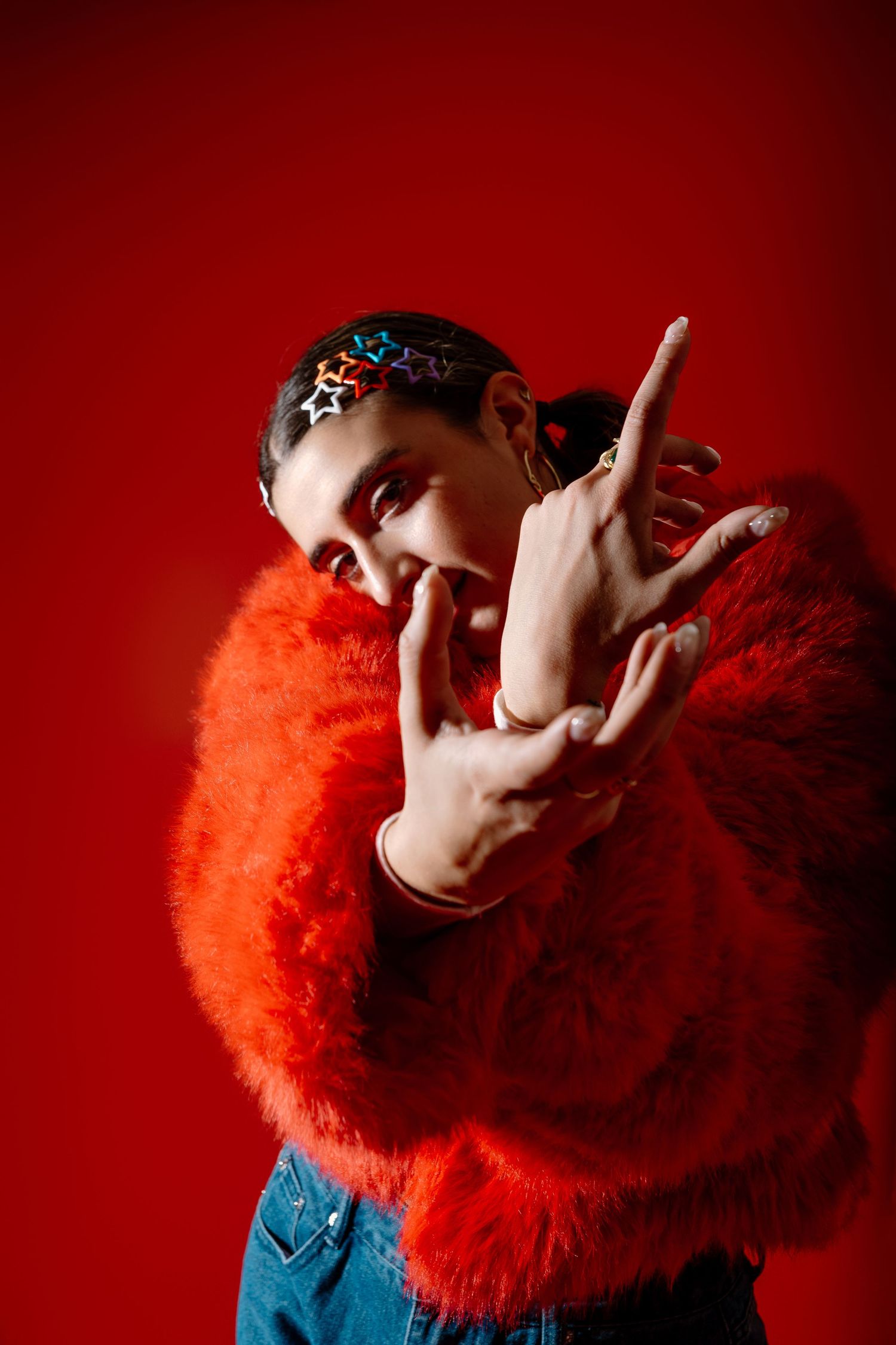 Person wearing bright orange fluffy sweater and blue jeans poses against red background with hand gestures.