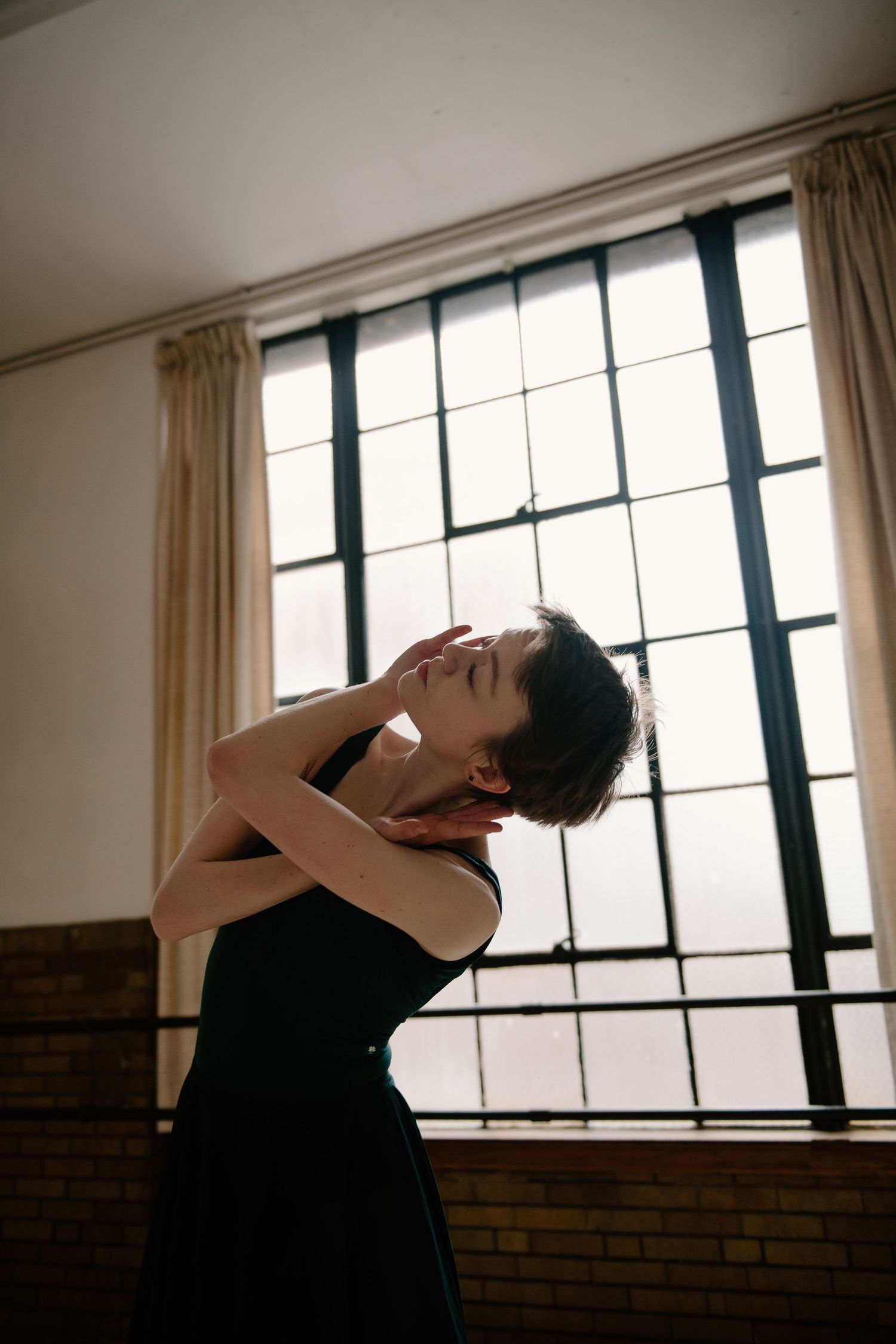 A dancer in a black dress poses dramatically near a large window with soft natural light streaming in.