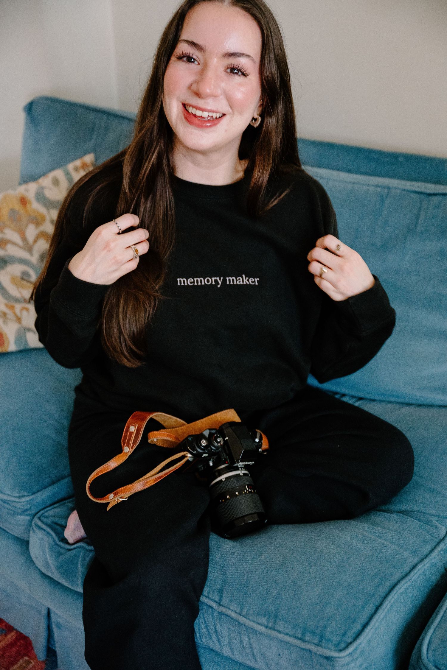 Smiling PHOTOGRAPHER woman in black memory maker sweatshirt sitting on blue sofa holding a DSLR camera with brown leather strap.