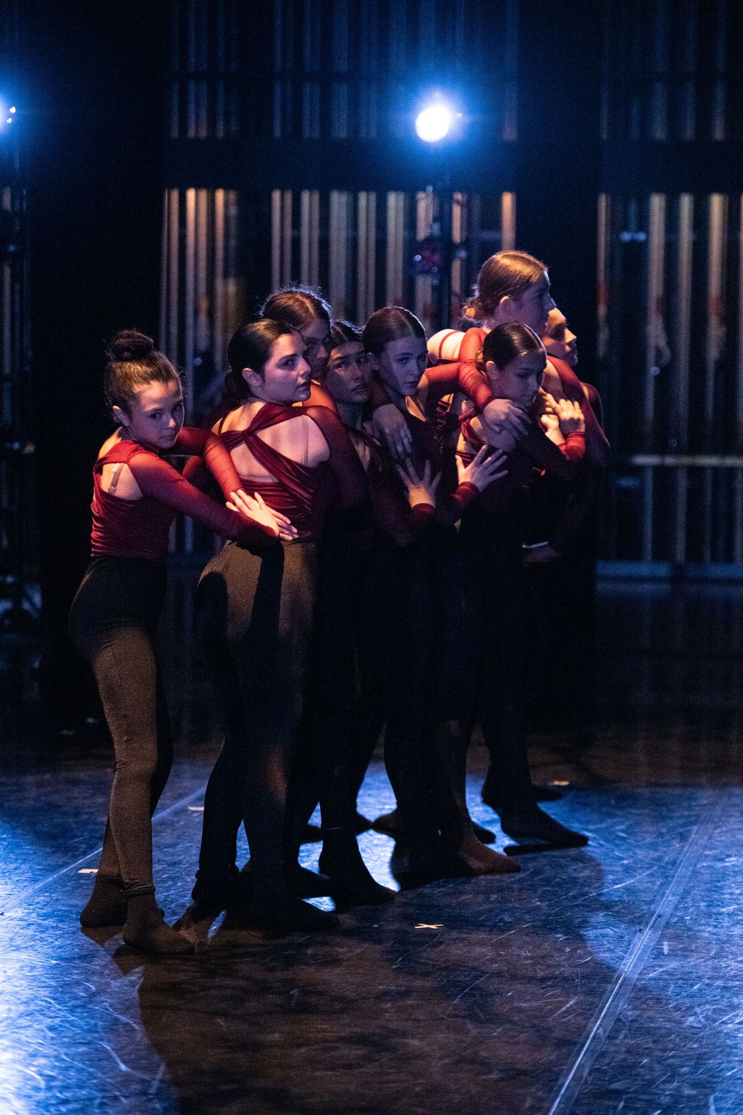 Dancers in red tops and black pants huddle together on a dimly lit stage during a dramatic performance.