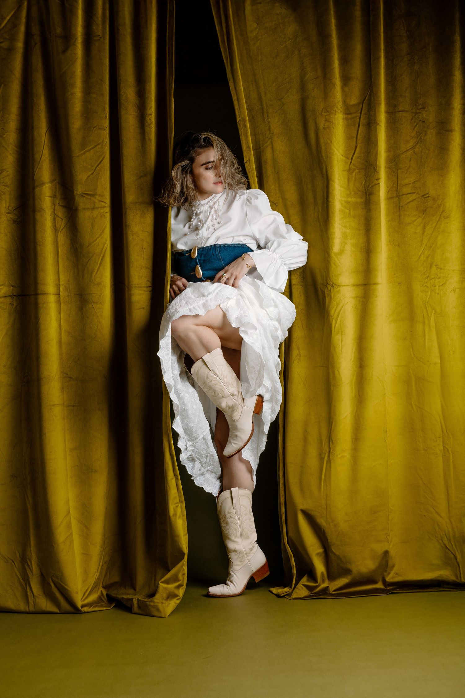 Woman in white ruffled dress, teal corset, and cream cowboy boots posing between golden velvet curtains.