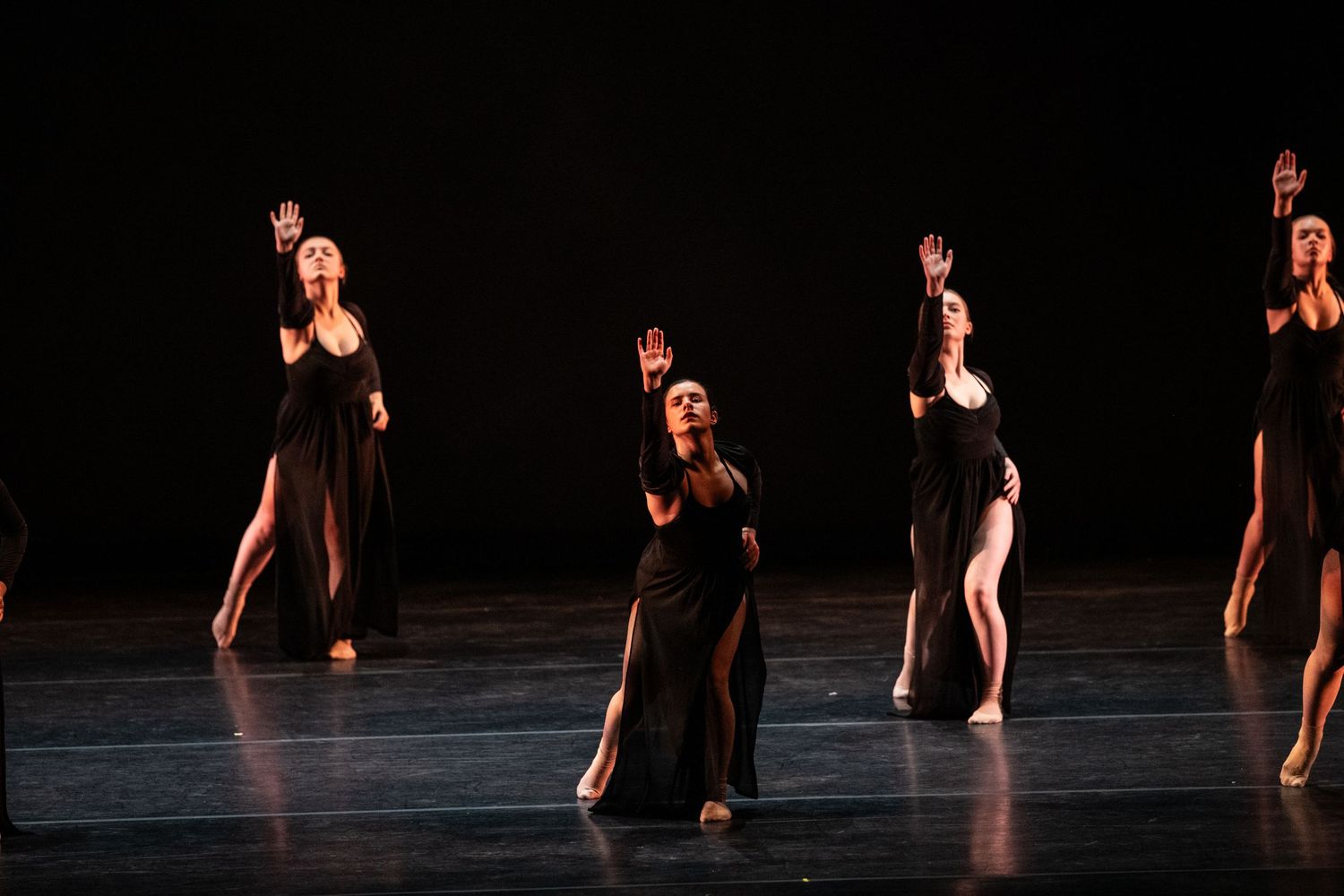 Dancers in black gowns perform on a dimly lit stage, arms raised gracefully in a dramatic contemporary dance performance.