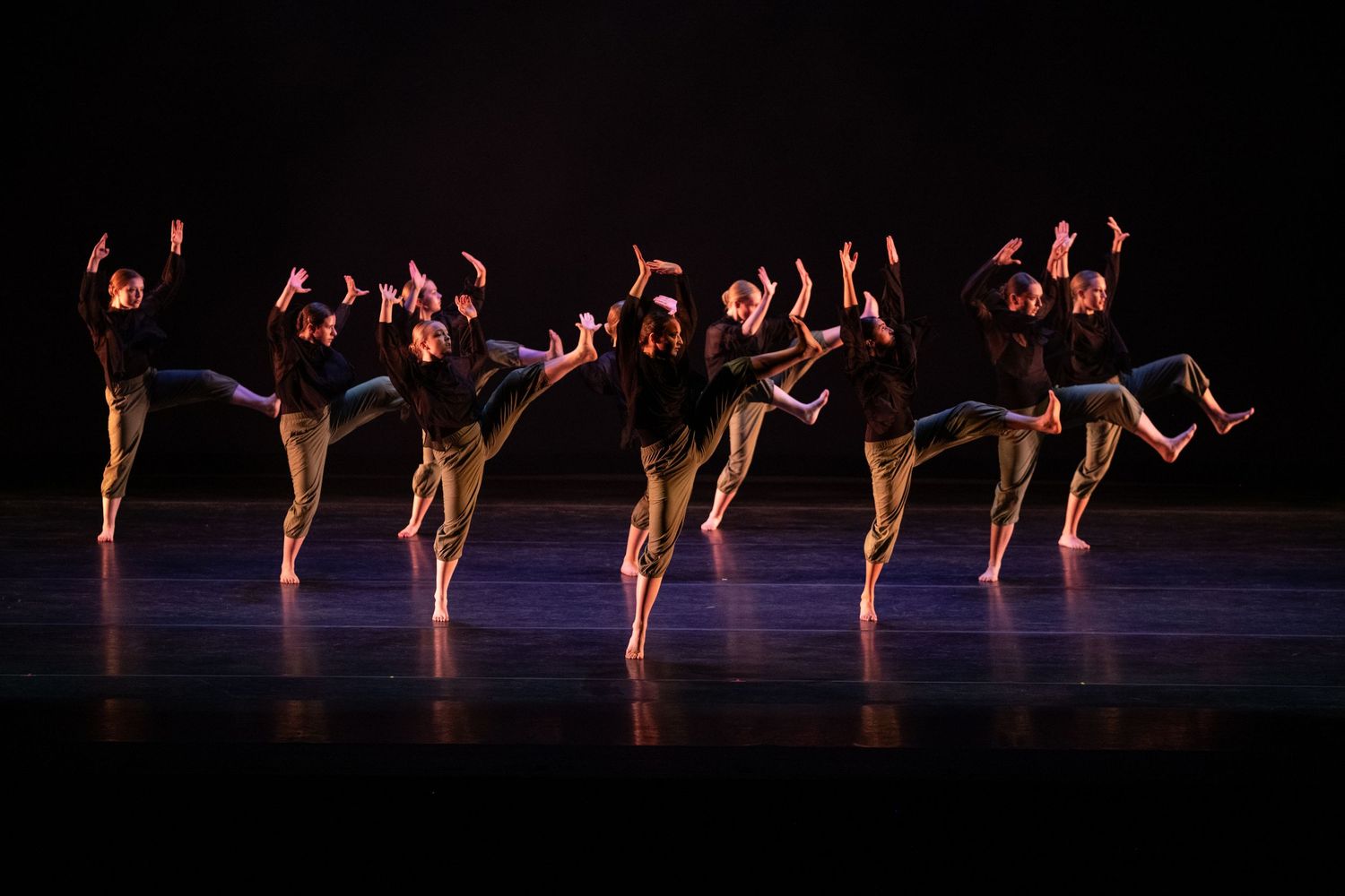 Dancers in black tops and tan pants perform synchronized contemporary dance moves on a dimly lit stage.