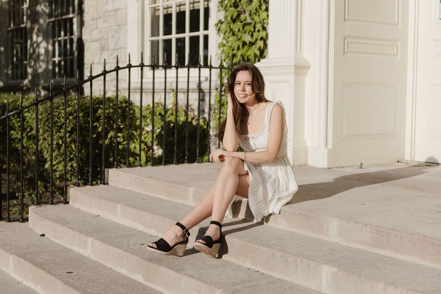 Woman in white dress poses gracefully on outdoor steps near elegant white columns and climbing vines.