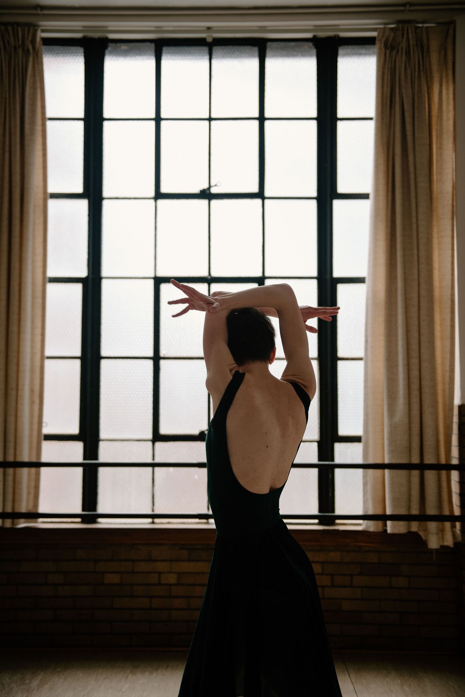 A silhouetted dancer in a black dress stretches gracefully against a bright window with curtains in a studio space.