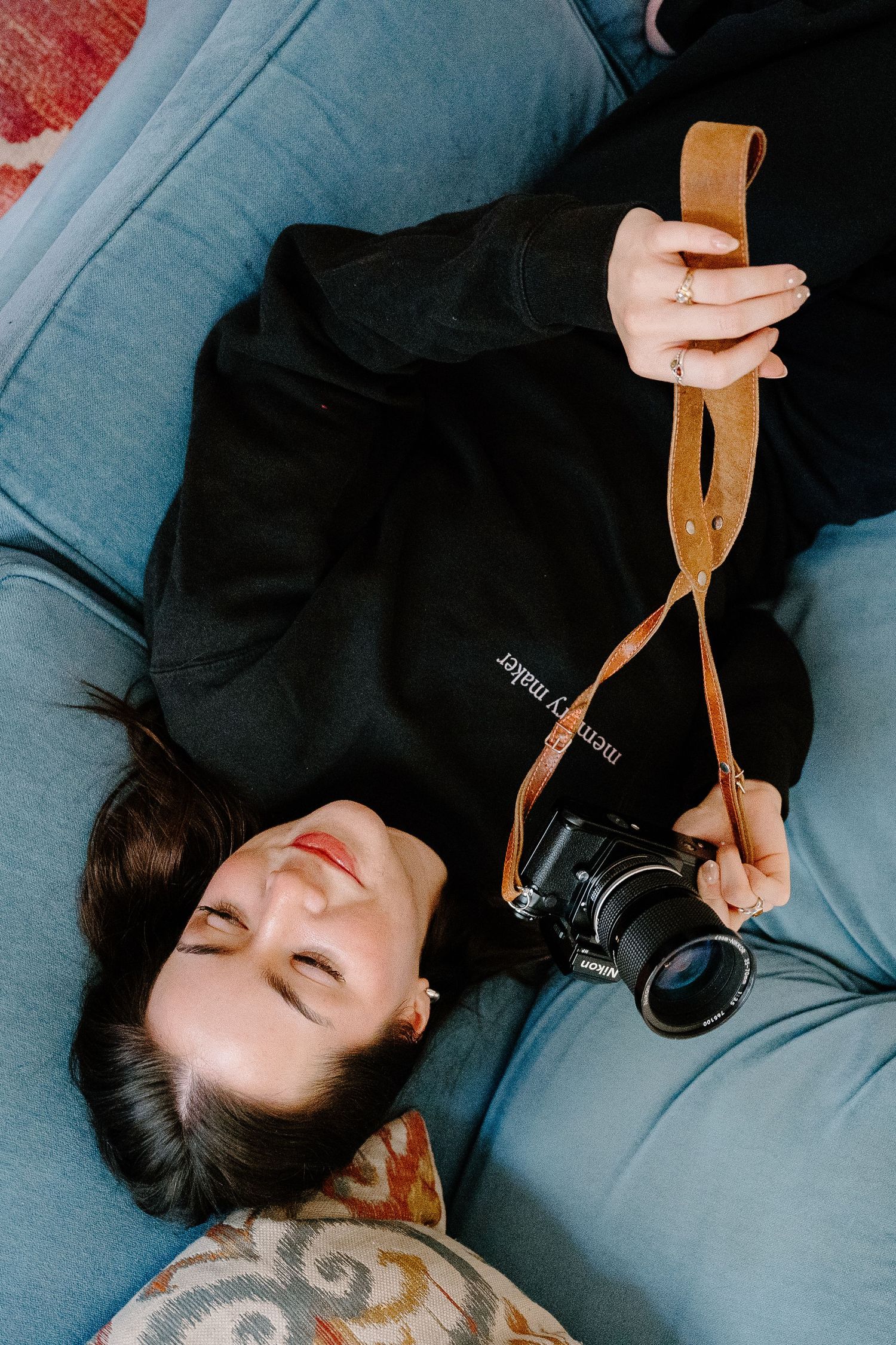 PHOTOGRAPHER in black hoodie lying on blue sofa holding a camera with leather strap, eyes closed, relaxed pose.