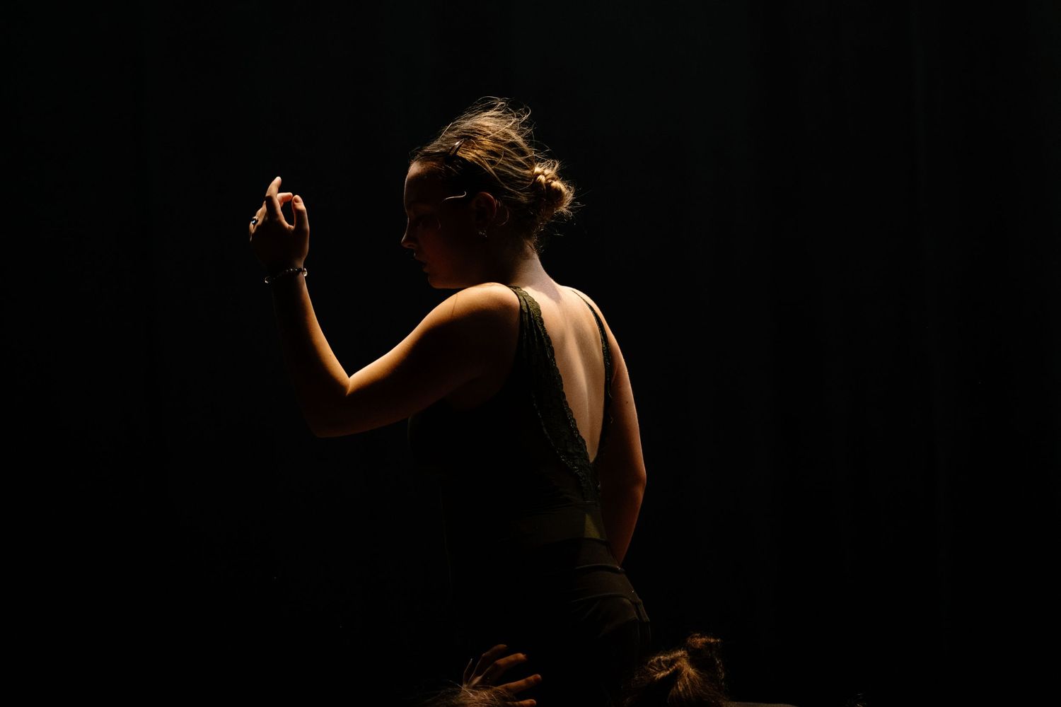 A woman dancer poses gracefully in dramatic side lighting against a dark background, wearing a sleeveless black outfit.