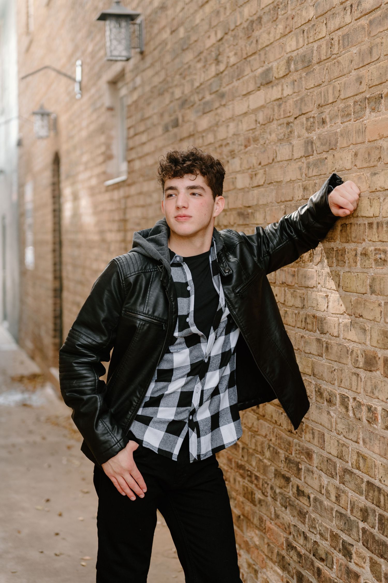 Person in black jacket and plaid shirt posing against brick wall backdrop.