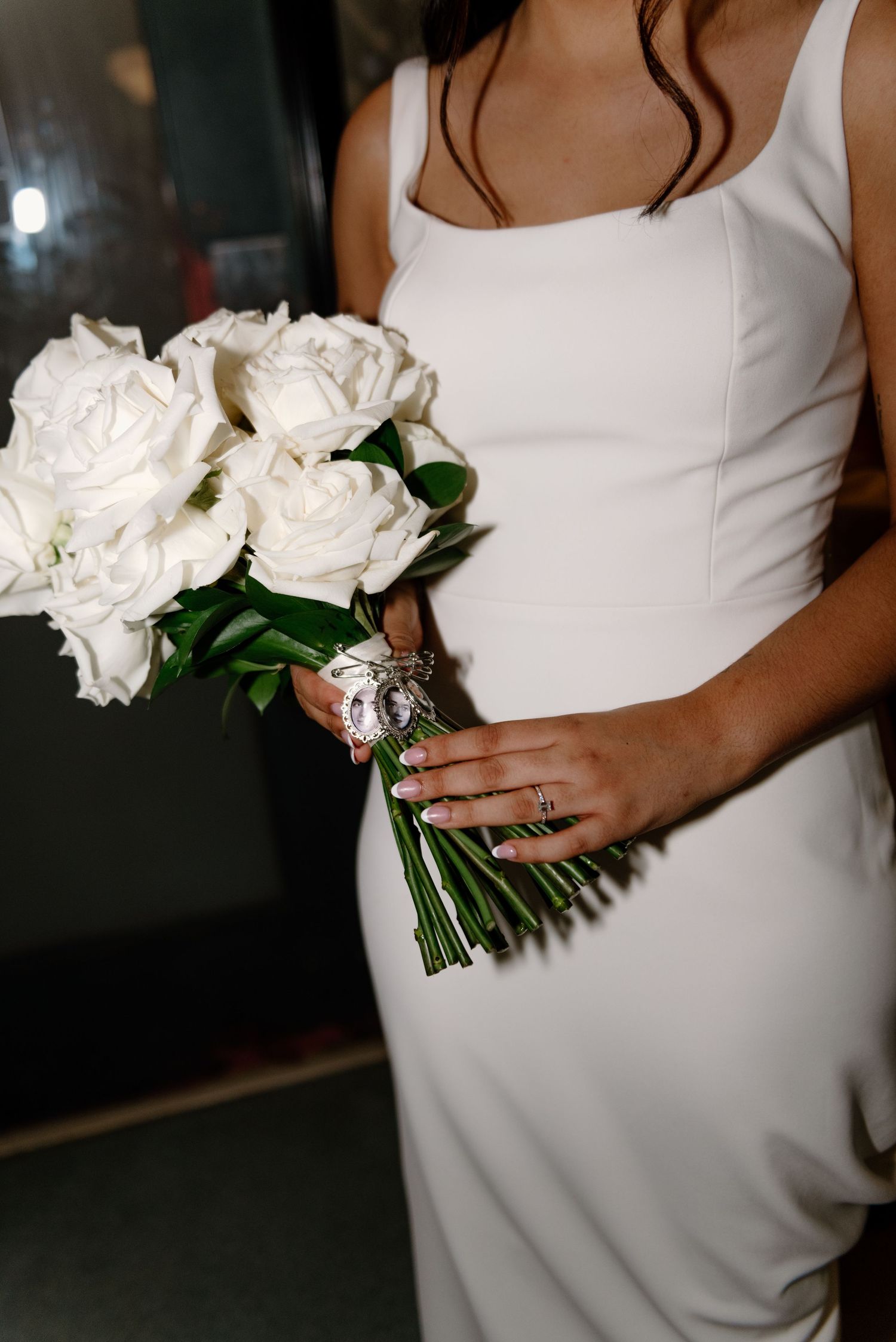 A white bridal bouquet with roses is held against a white wedding dress in an elegant indoor setting.