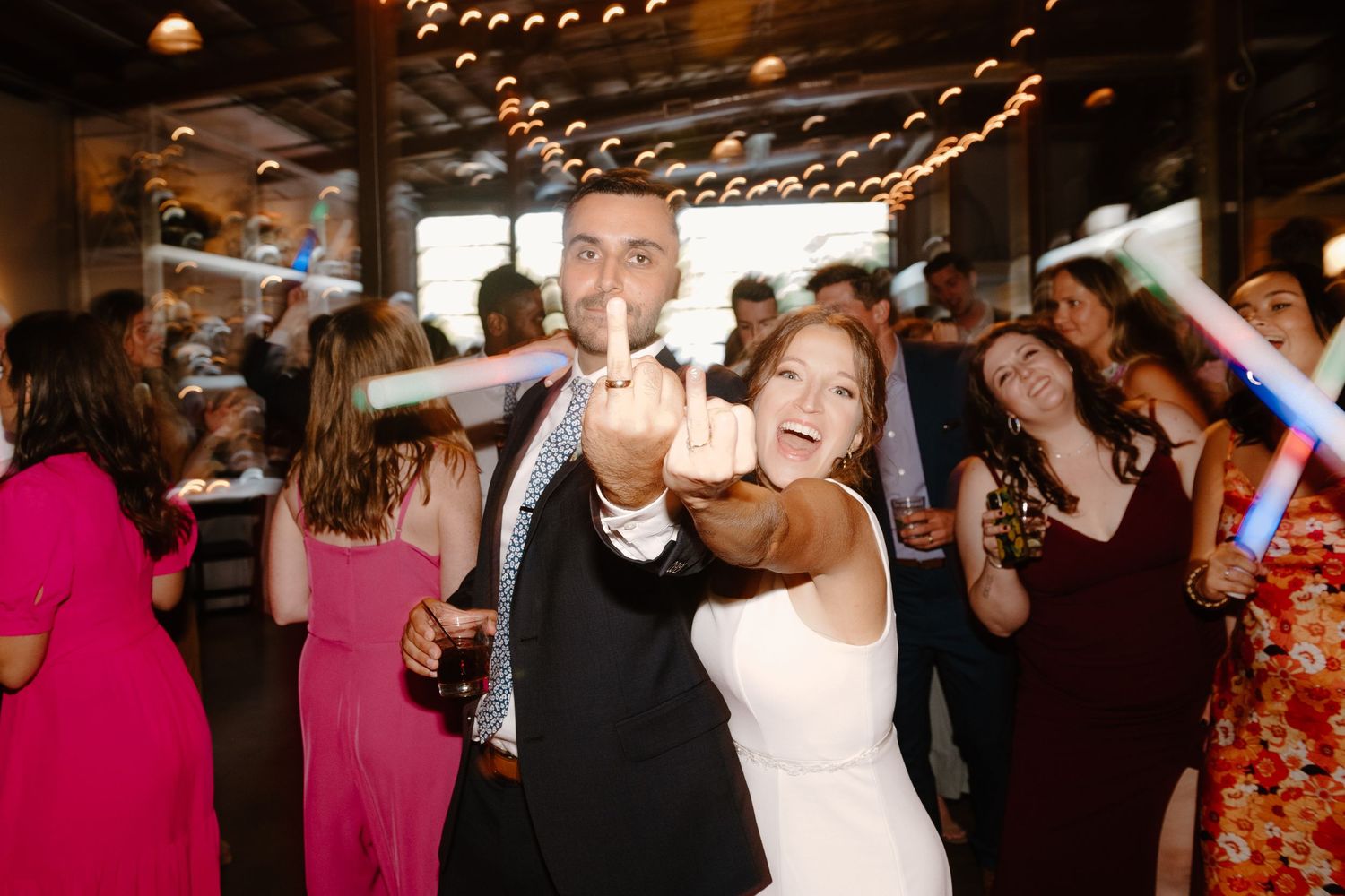 Wedding guests dancing and celebrating on a dimly lit dance floor under string lights during an evening reception.