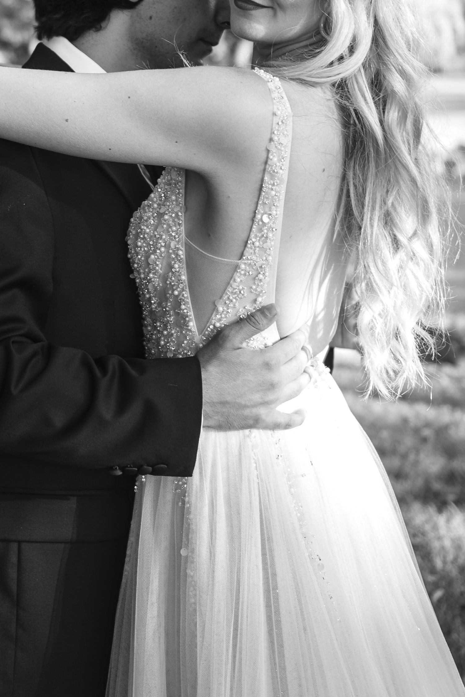 Black and white photo of a bride and groom embracing, showcasing her beaded open-back wedding gown and flowing hair.