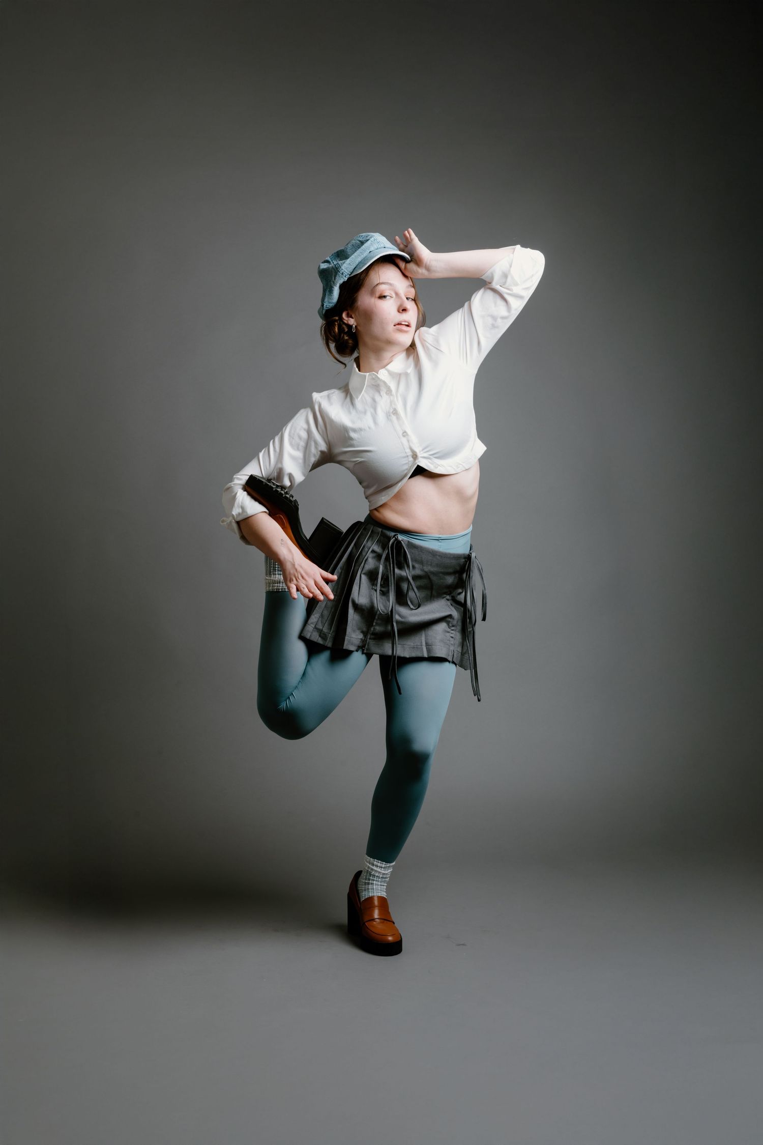 A person in blue tights and a cropped white top strikes a dynamic dance pose against a gray studio backdrop.