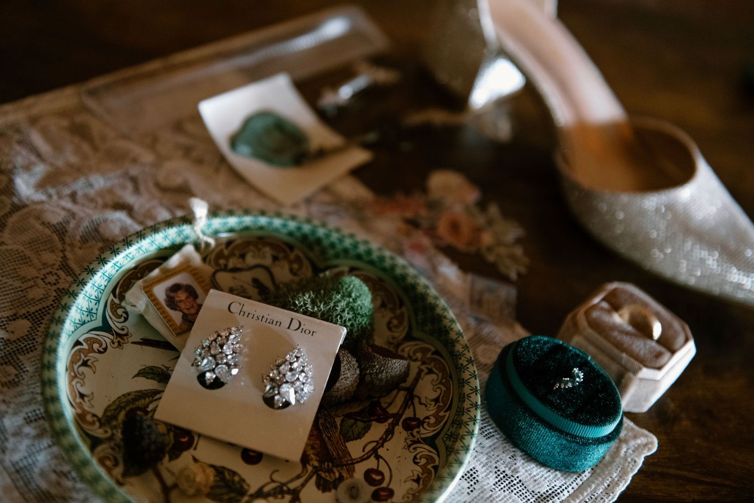 Christian Dior crystal earrings displayed in a green dish alongside a ring box and glittery bridal heels on a wooden table.