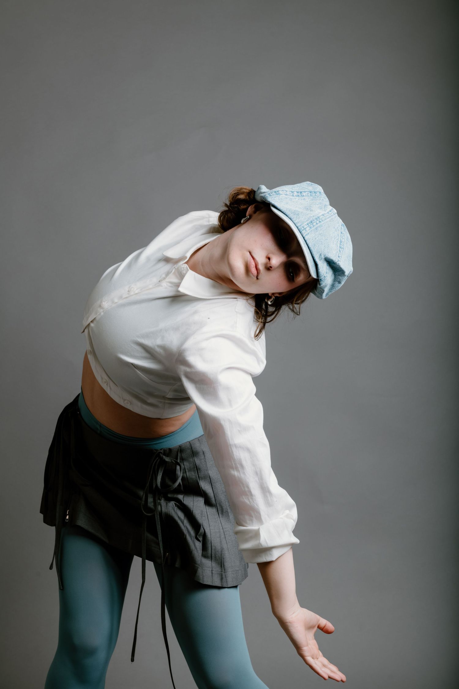 Woman in light blue cap, white cropped shirt and dark shorts strikes a dance pose against gray backdrop.