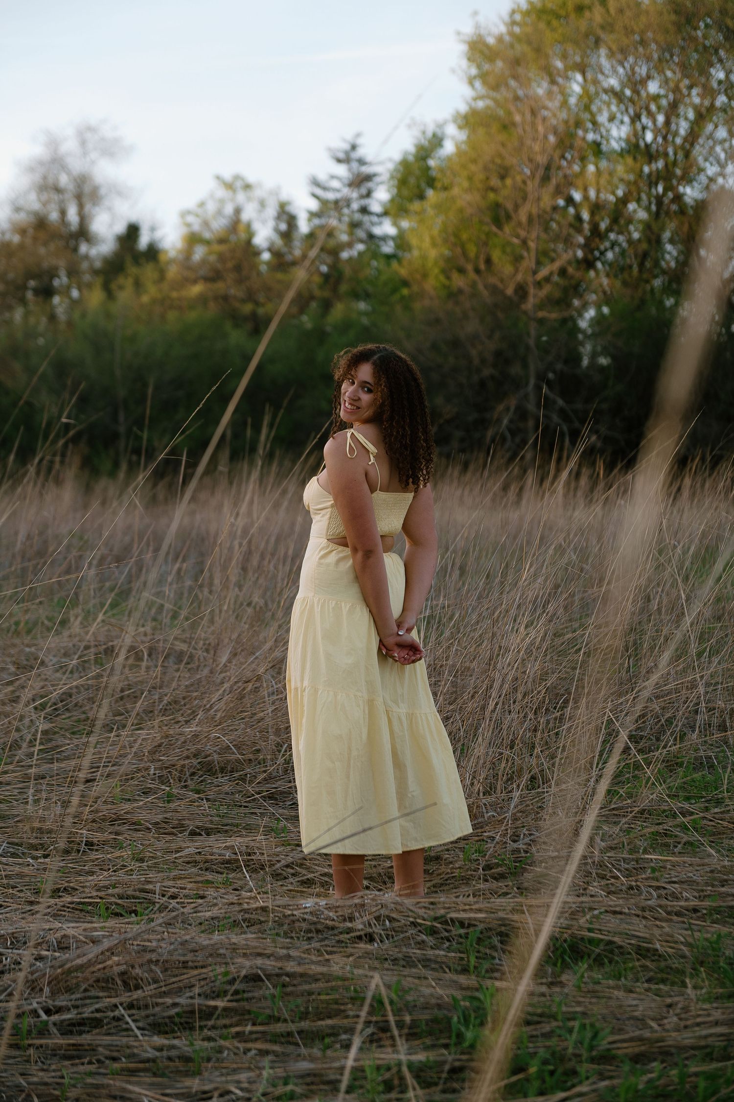 Smiling woman in a flowing pale yellow sundress stands barefoot in a tall dry grass field, glancing over her shoulder with trees behind.