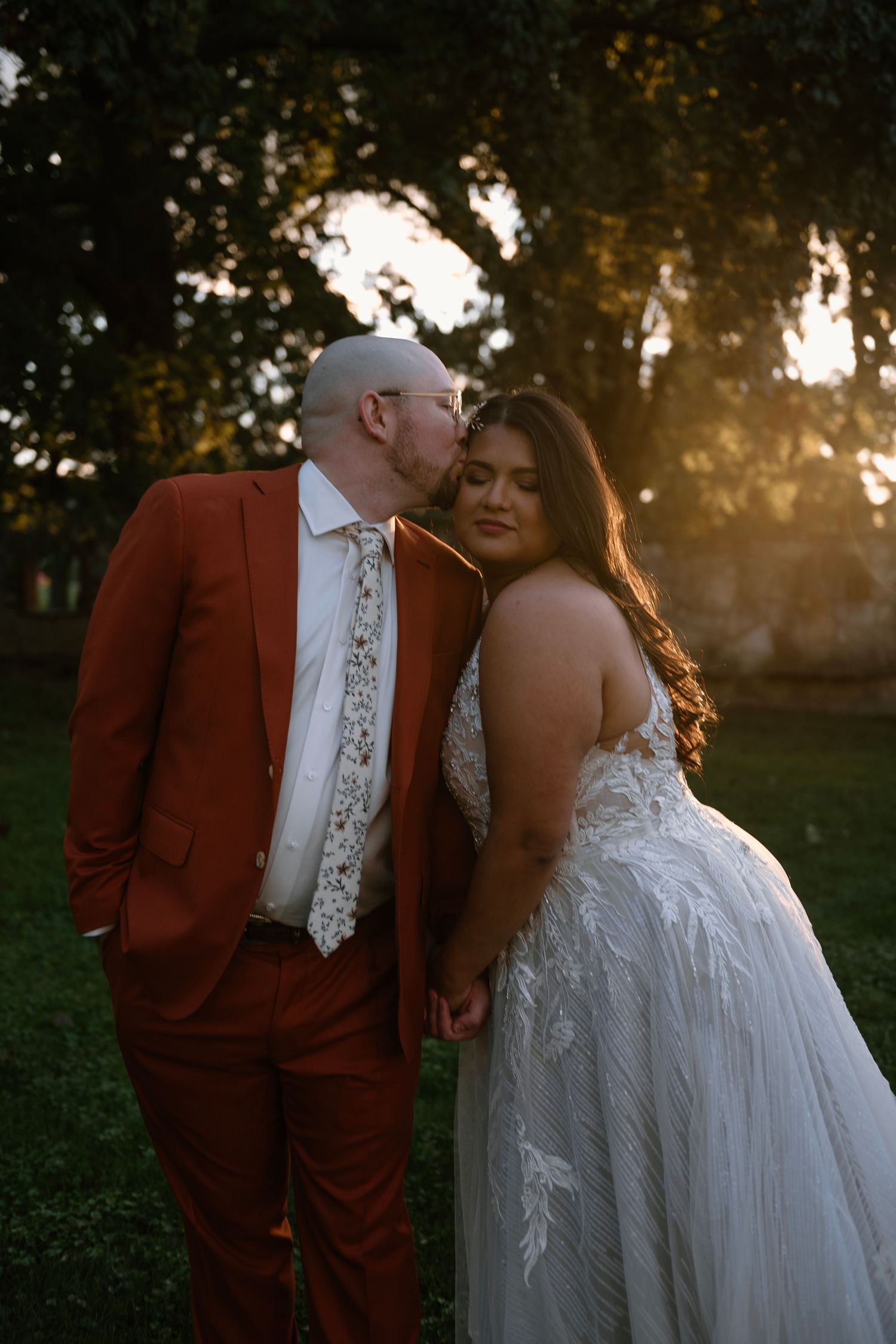 Groom in rust suit kisses bride in lace wedding gown during golden hour outdoors.