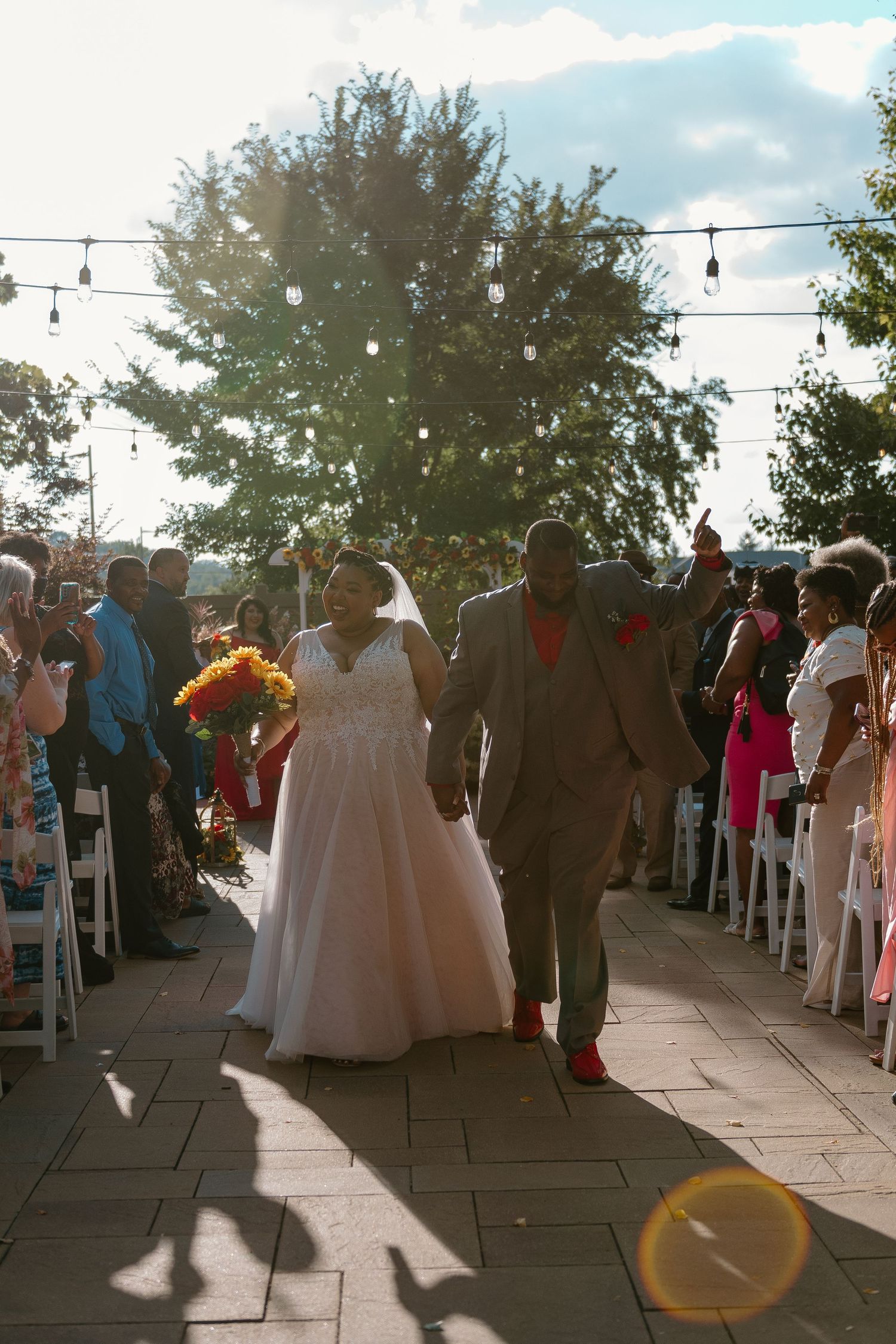 A couple walks down an outdoor aisle at sunset while guests look on from white chairs under string lights and trees.