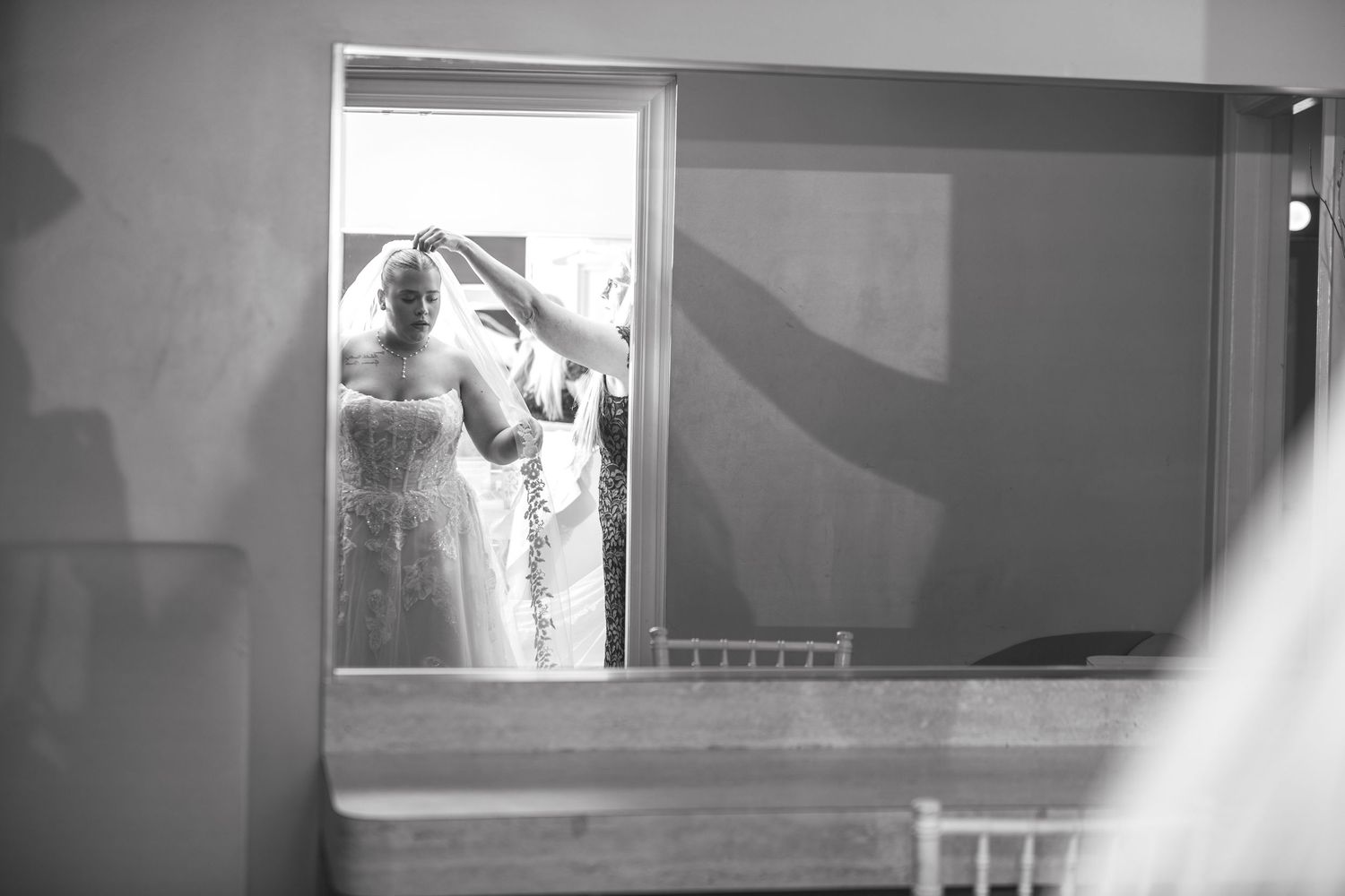 Black and white photo of a bride adjusting her veil in a mirror reflection during wedding preparations.