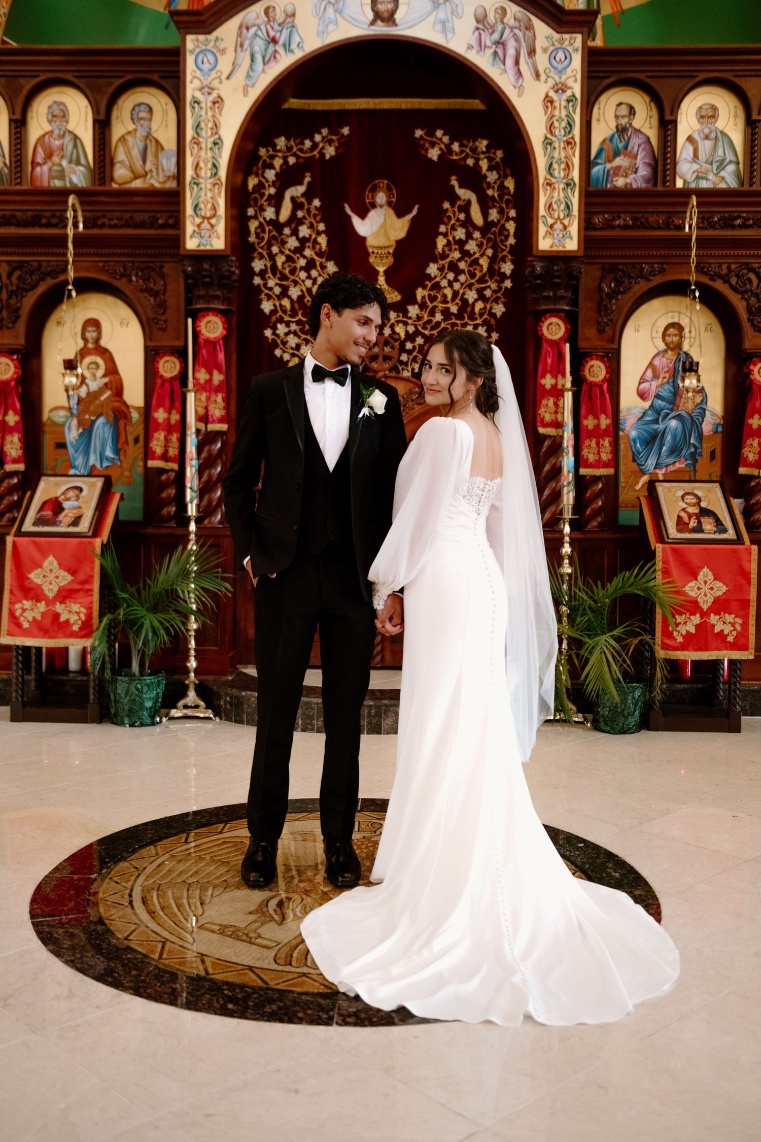 A couple poses in formal wedding attire inside an ornate Orthodox church with religious icons and artwork on display.