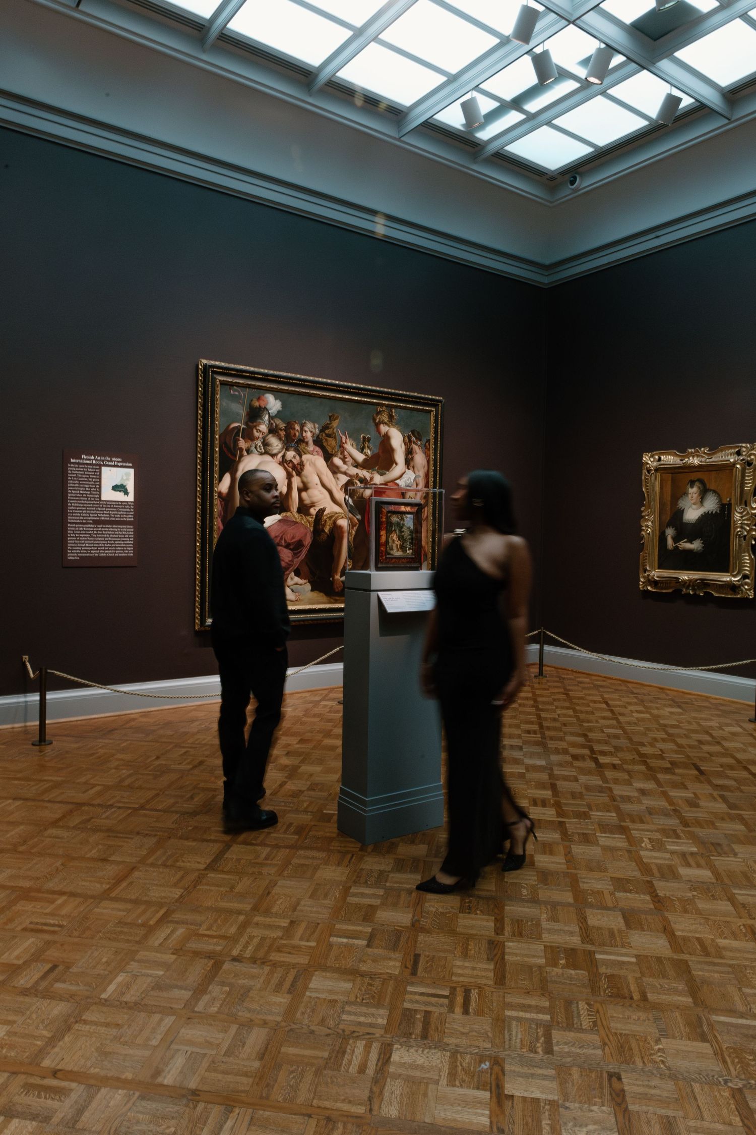 Two visitors admire classical paintings in a dimly lit museum gallery with parquet floors and skylight.