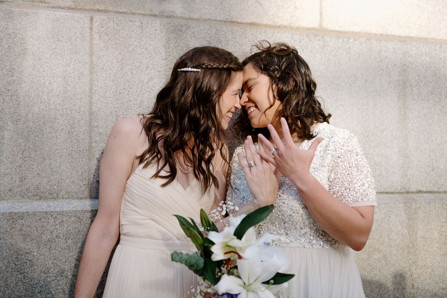 Two brides in white wedding dresses share an intimate moment while holding a white floral bouquet against a stone wall.