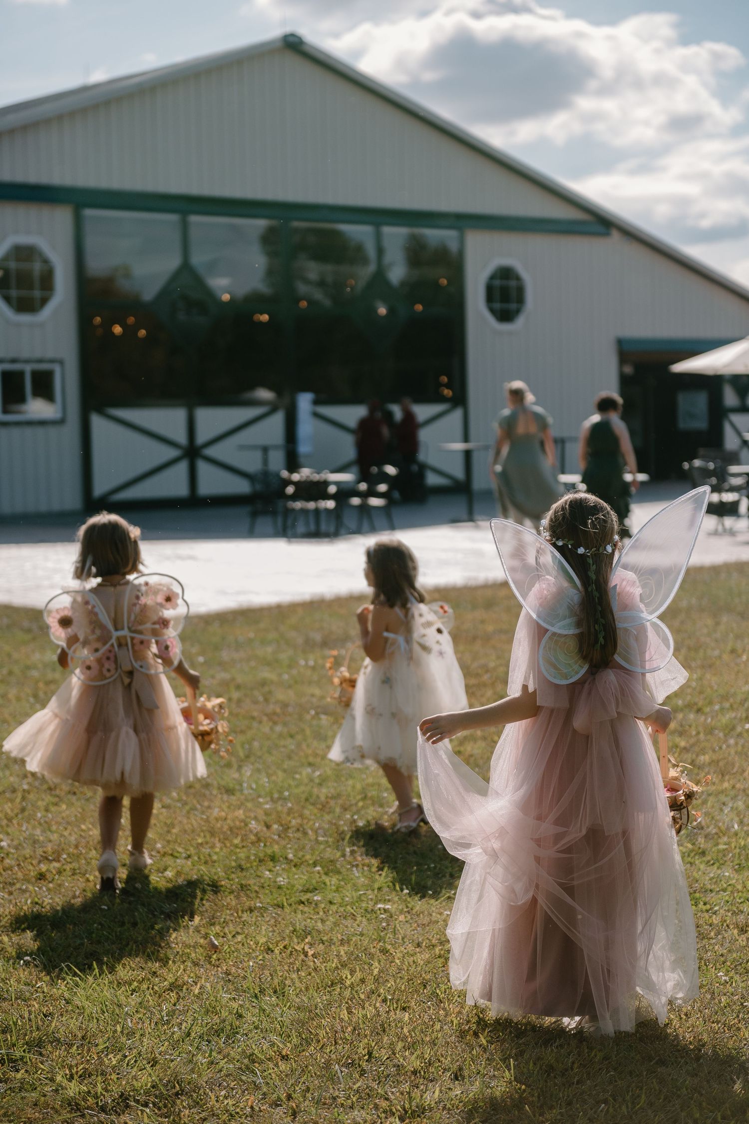 Three flower girls in pink tulle dresses and fairy wings walk across a lawn towards a white barn wedding venue.