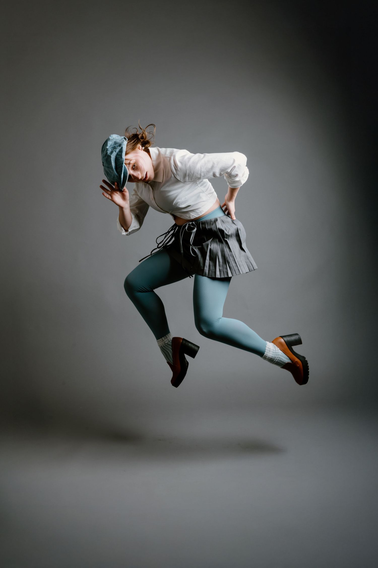 Dynamic studio shot capturing a dancer in mid-leap wearing a white button-down shirt, gray skirt and blue tights.