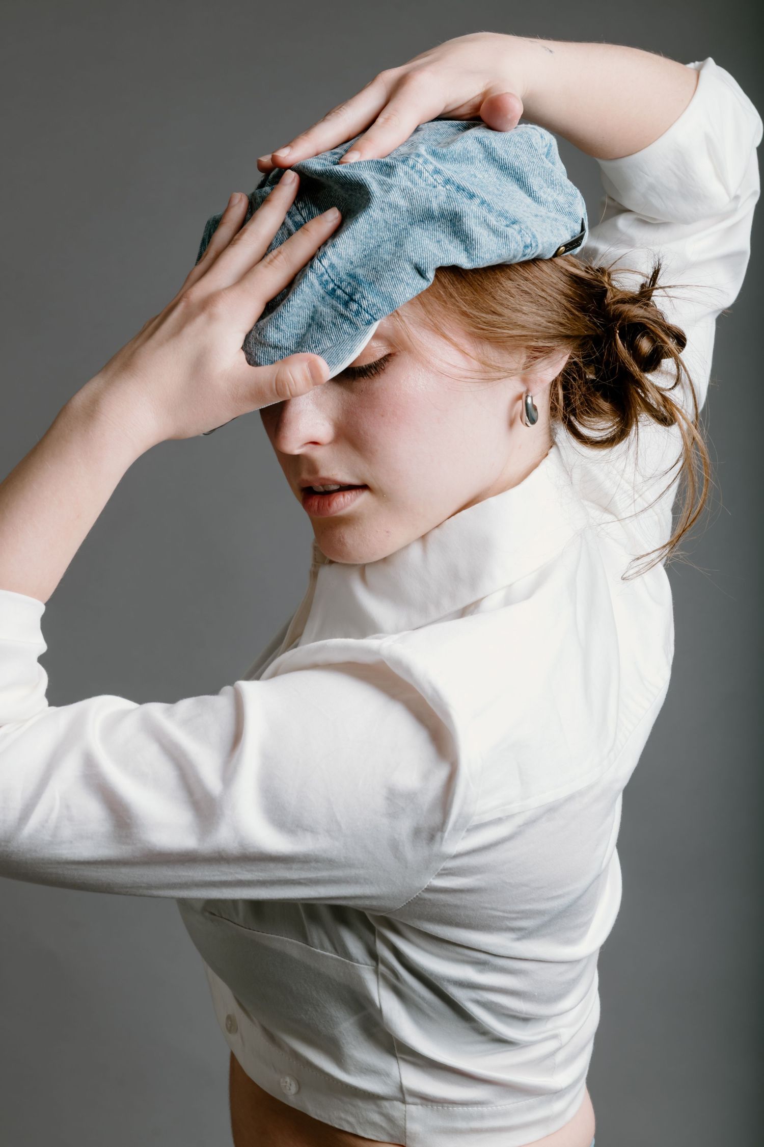 Person in white long-sleeved top holds blue fabric to forehead in dramatic artistic pose against grey background.