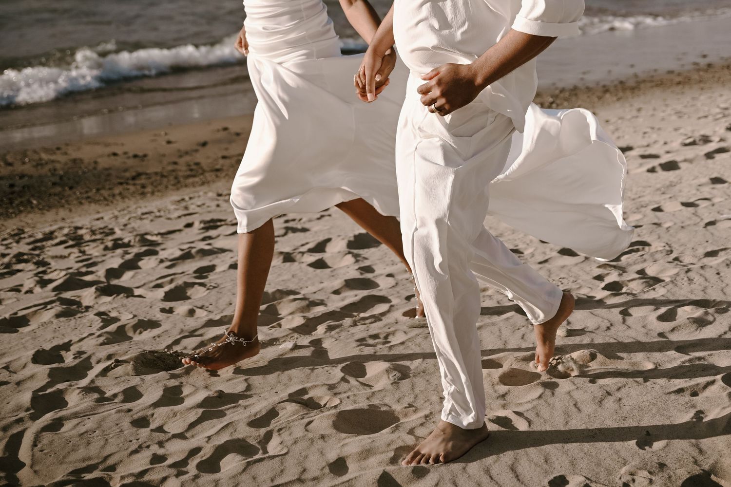 Couple in flowing white outfits walking barefoot along a sandy beach at sunset.