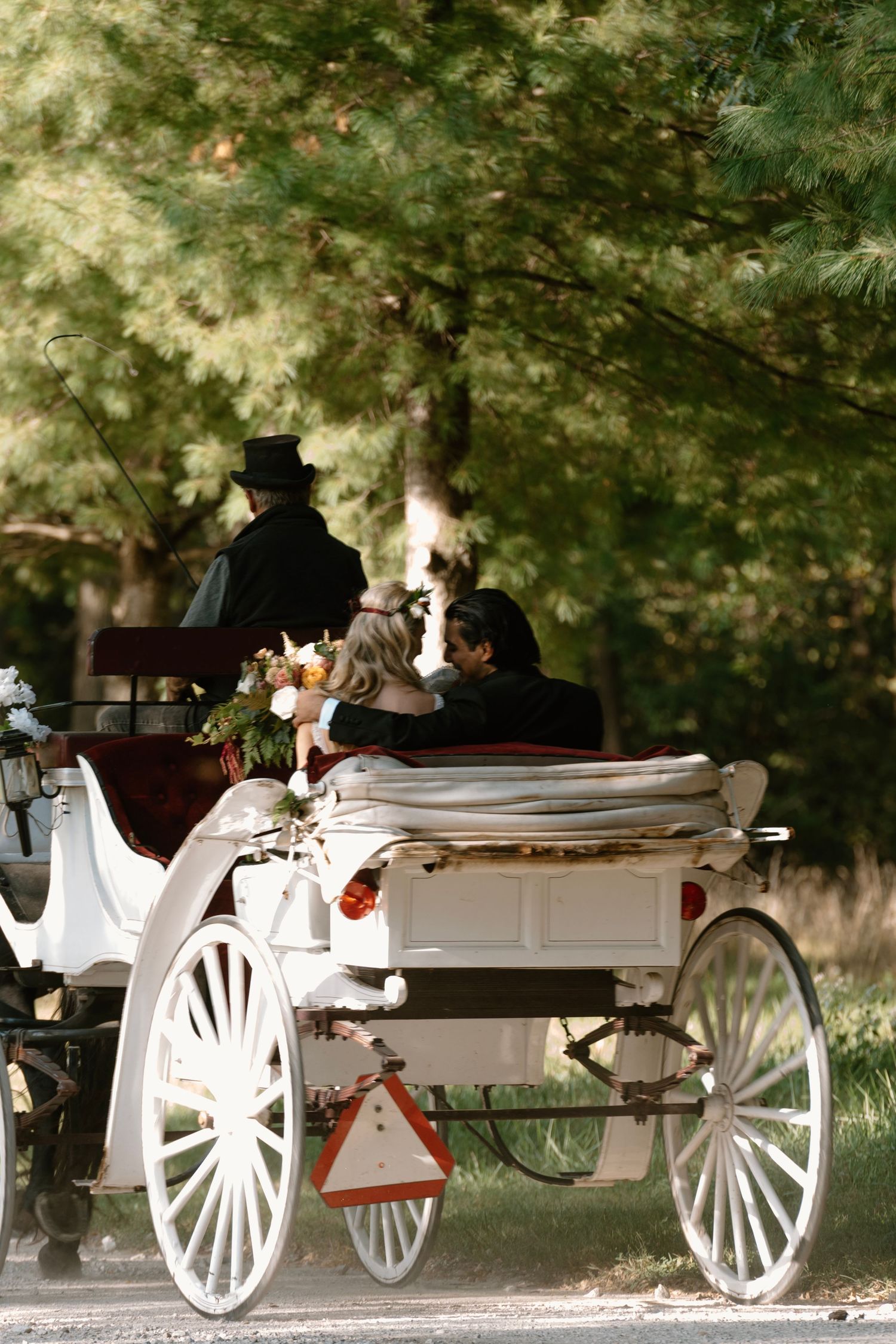 Couple riding in a white horse-drawn carriage decorated with flowers, guided by a coachman through a tree-lined path.