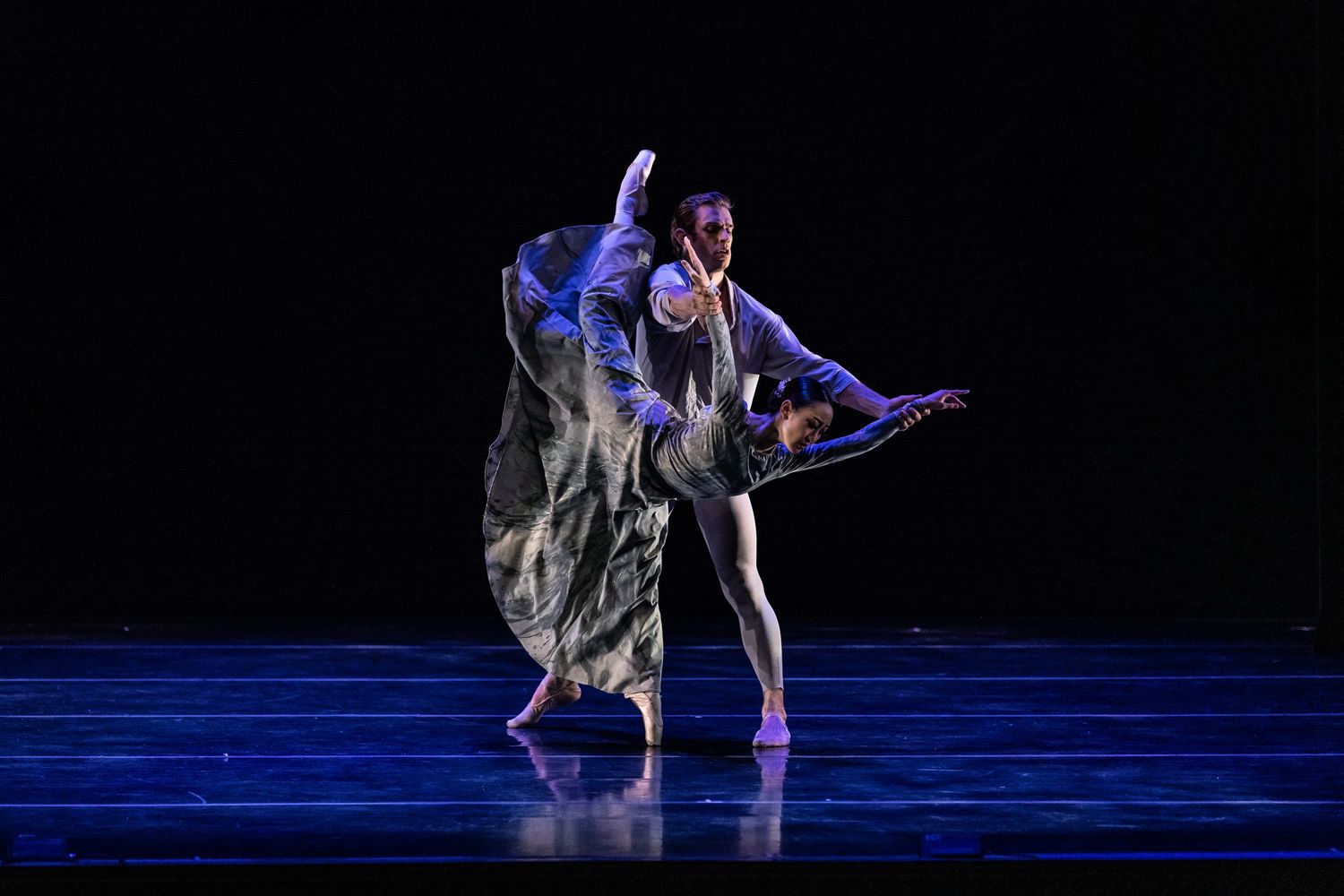 Dancers perform contemporary ballet movements on a dark stage illuminated by blue lighting.