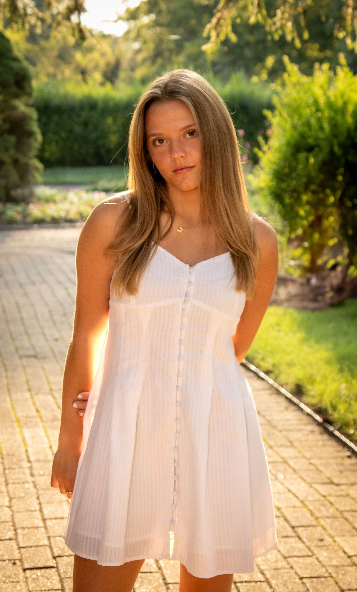 Person in white summer dress poses on brick pathway in garden setting.