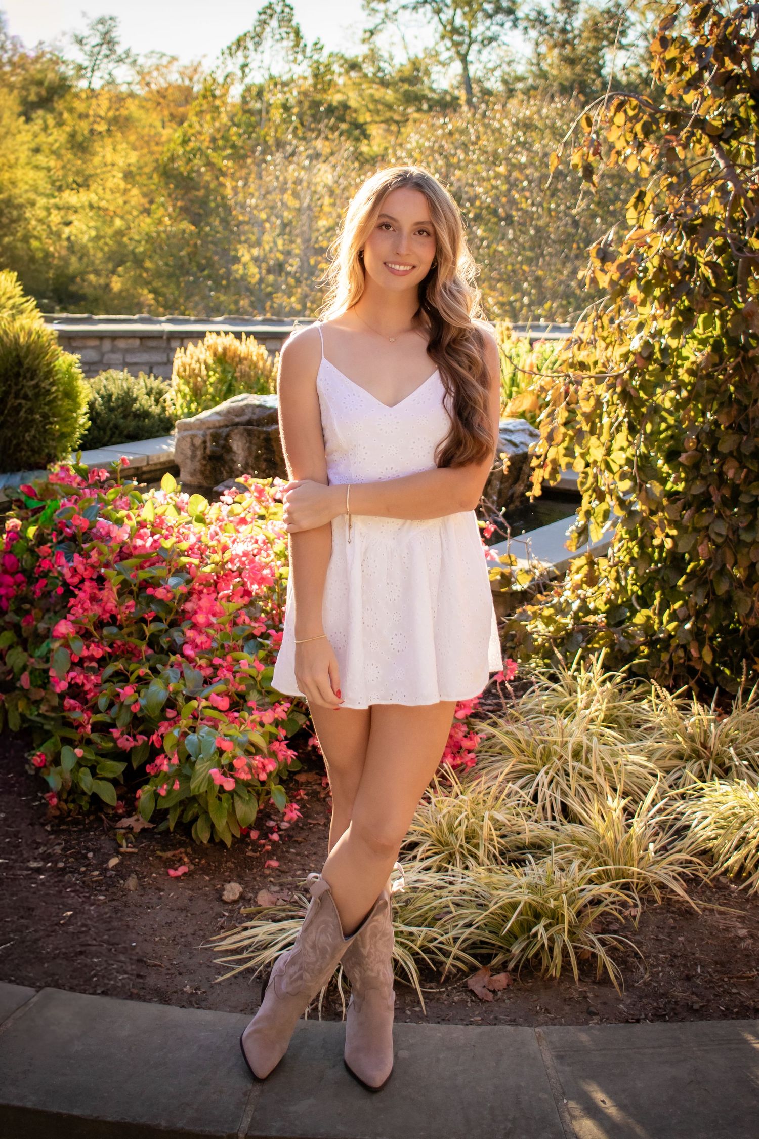 Woman in white dress poses in garden with pink flowers and autumn foliage creating a romantic outdoor setting.
