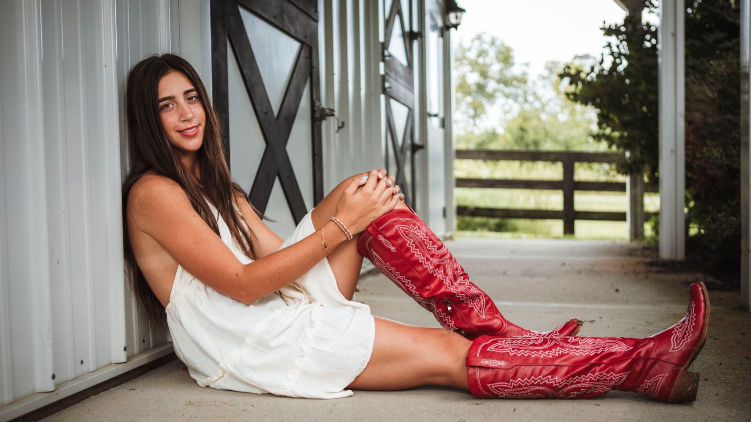 Young woman in white dress and red cowboy boots sitting against a barn wall outdoors.