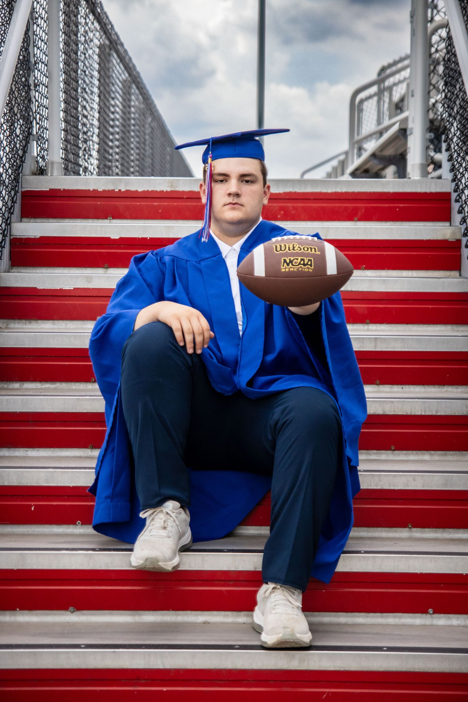 Young man in blue graduation cap and gown sits on red stadium steps holding a Wilson NCAA football.