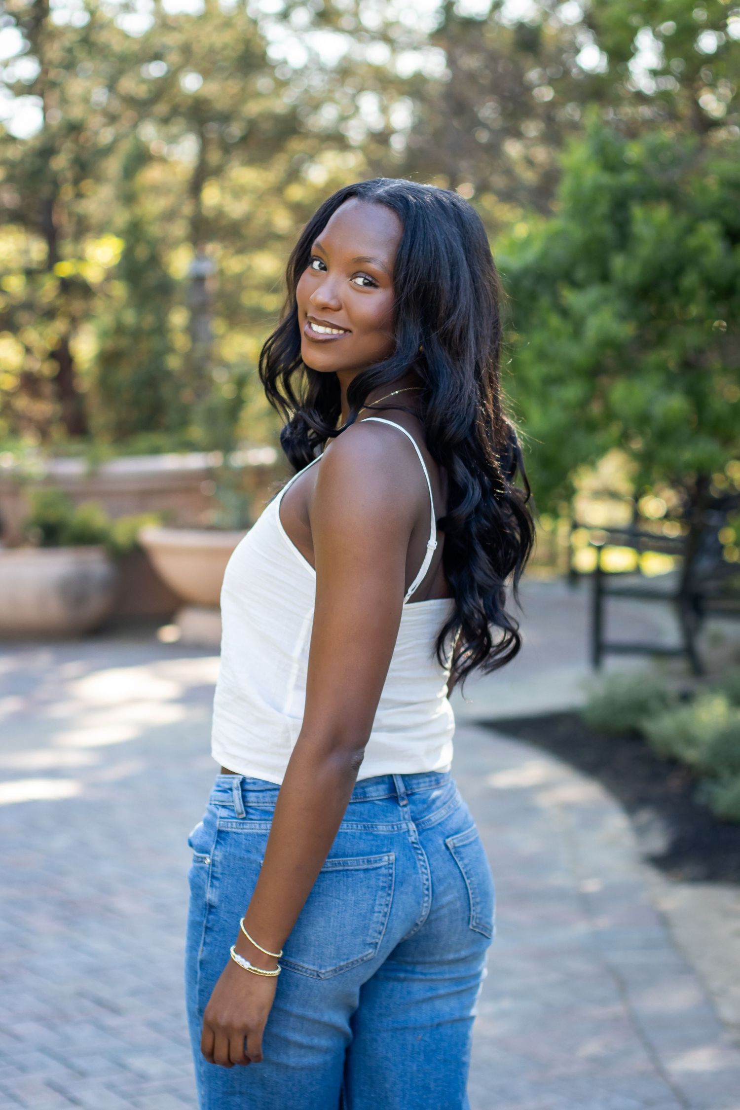 Smiling young Black woman in white tank top and blue jeans posing outdoors in a sunny park setting.