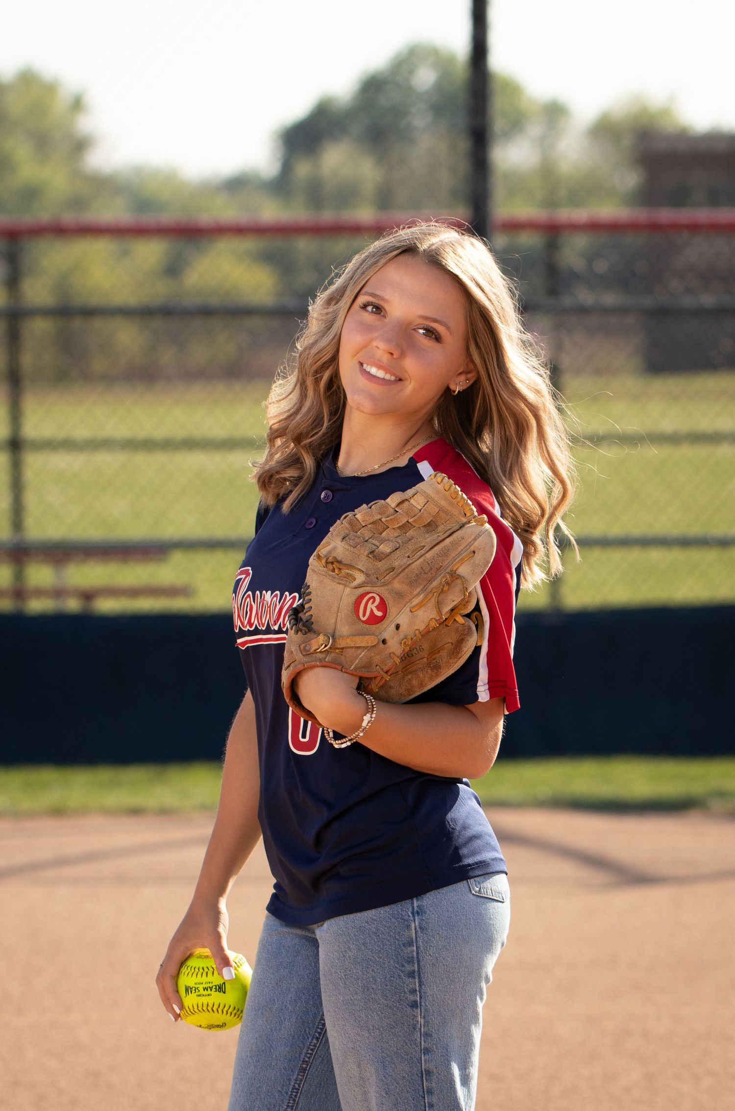 Smiling female softball player in navy jersey holding Rawlings glove and yellow softball on outdoor field.