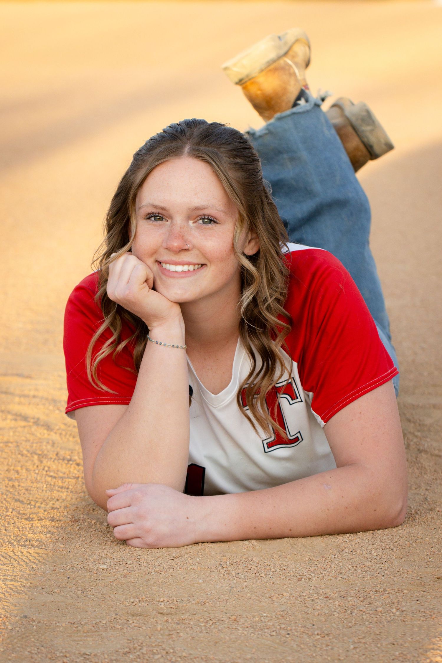 Smiling teenage girl in red jersey lying on sandy ground, resting chin on hand, wearing jeans and boots.