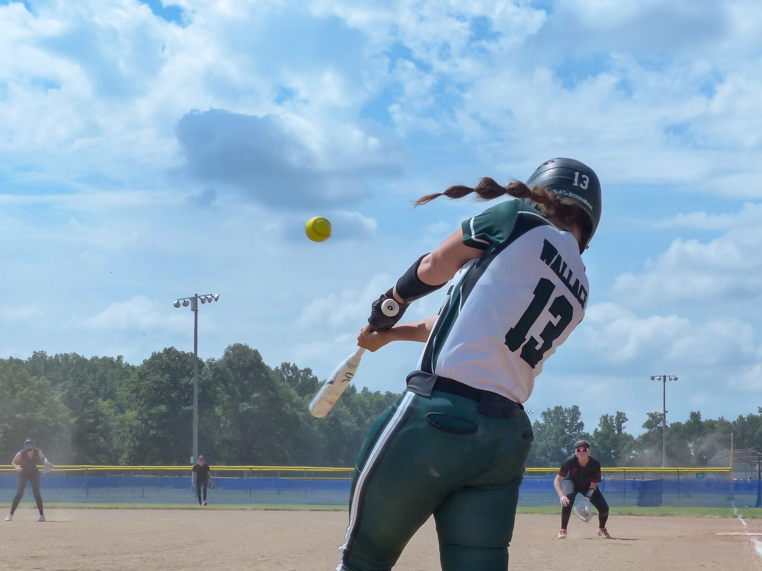 A softball player in green uniform swings their bat during a game on a sunny day with blue skies.