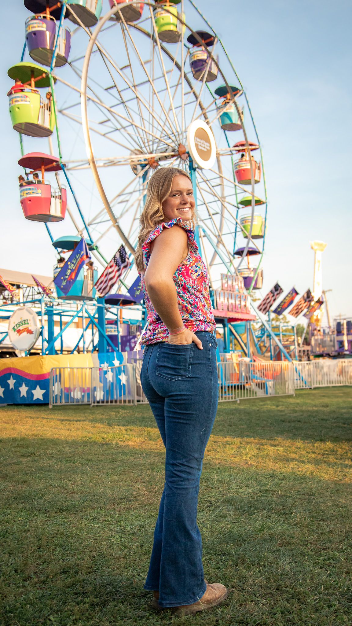 A person in a floral top and jeans poses in front of a colorful ferris wheel at a carnival fairground.