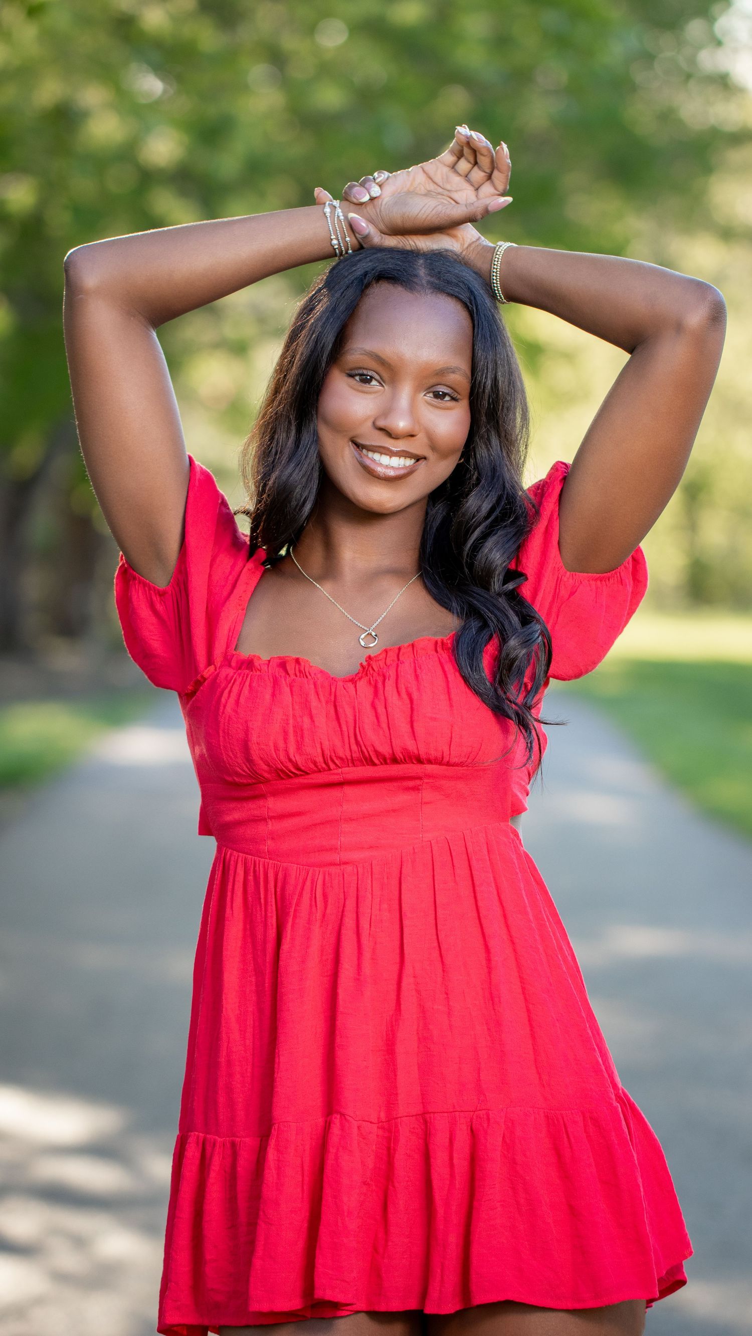 Smiling young woman in red mini dress poses outdoors on a tree-lined path with hands raised above head.