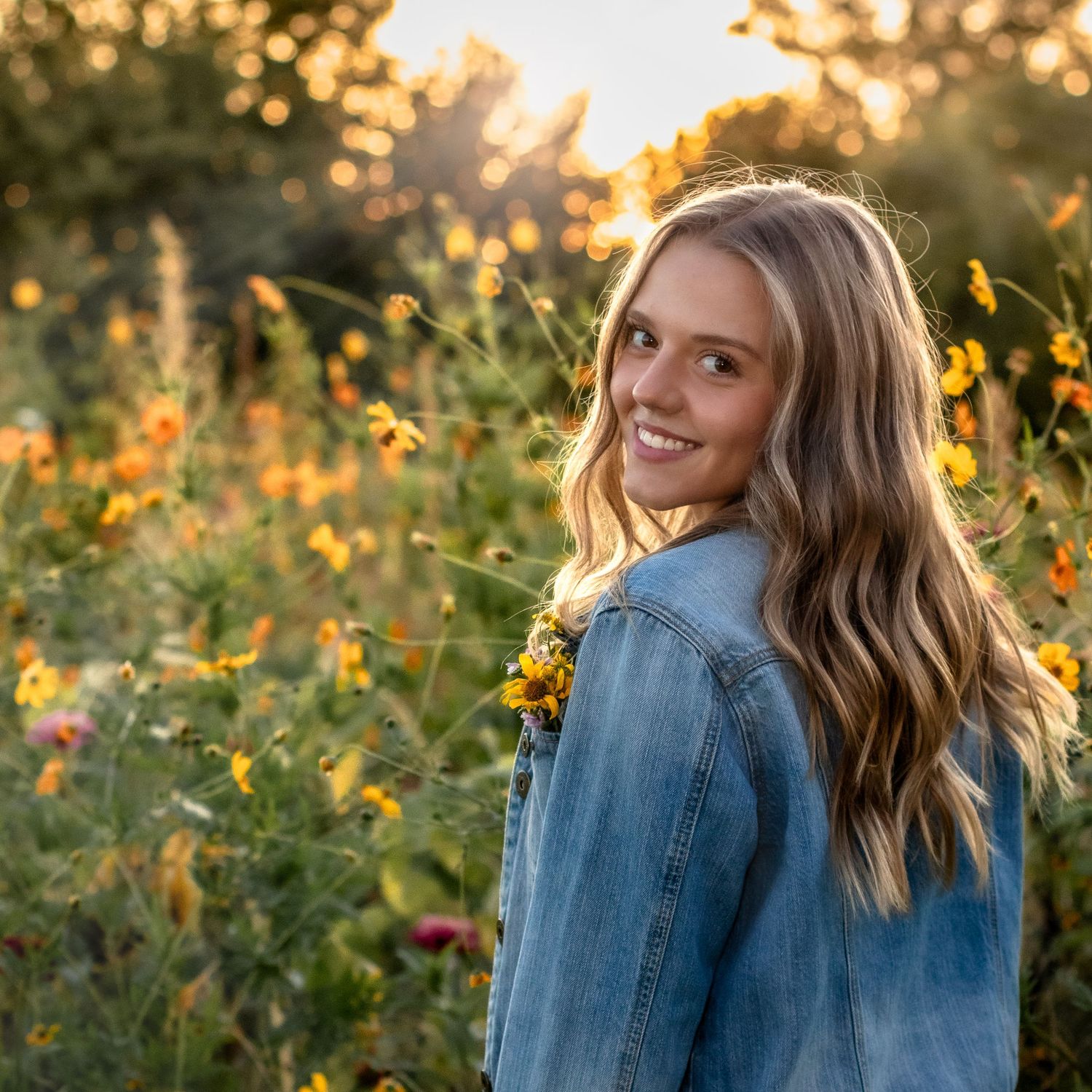 Young woman in denim jacket smiling over shoulder in golden wildflower field at sunset.