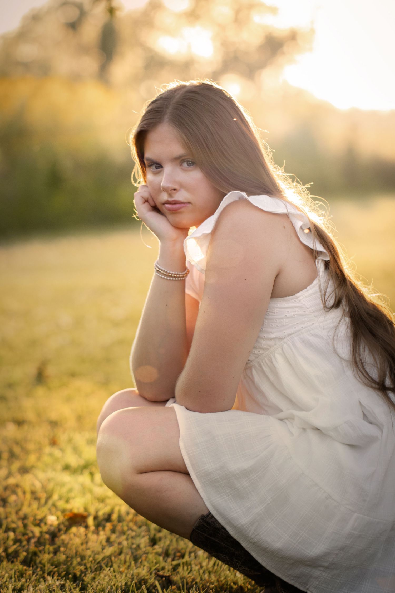 Young woman in white dress sitting on grass in golden sunset light, looking over shoulder with contemplative expression.