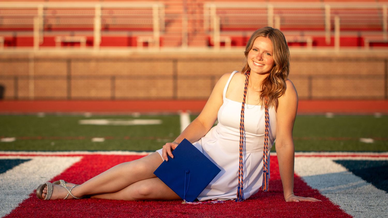 Smiling graduate in white dress holds blue cap on red turf football field for senior portrait photo.