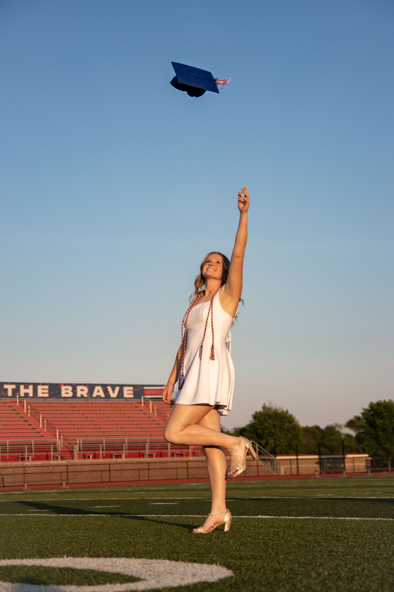 A young woman in a white dress tosses her blue graduation cap into the air on a football field.