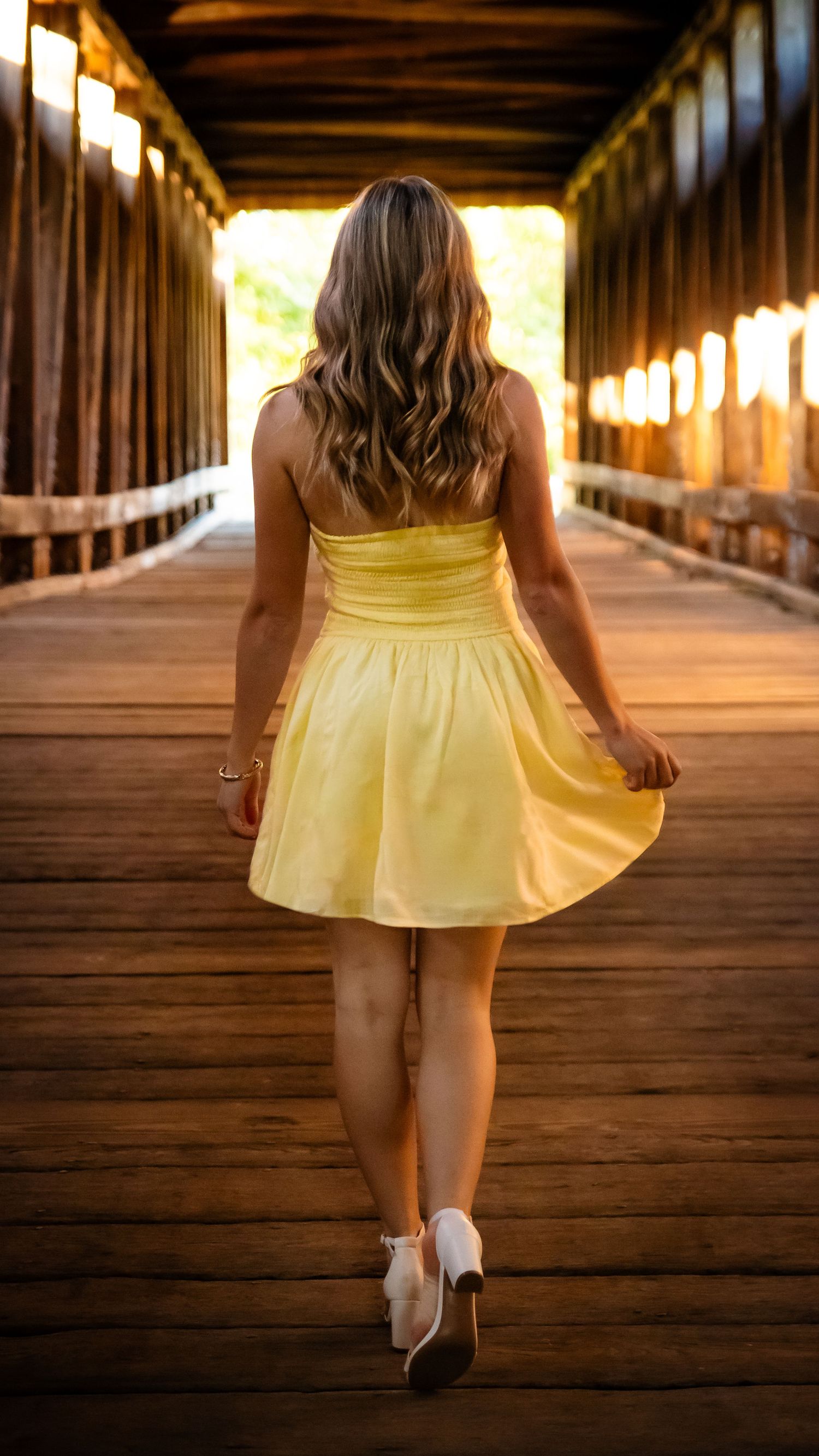 A young child in a yellow dress walks through a covered wooden bridge on a sunny day.