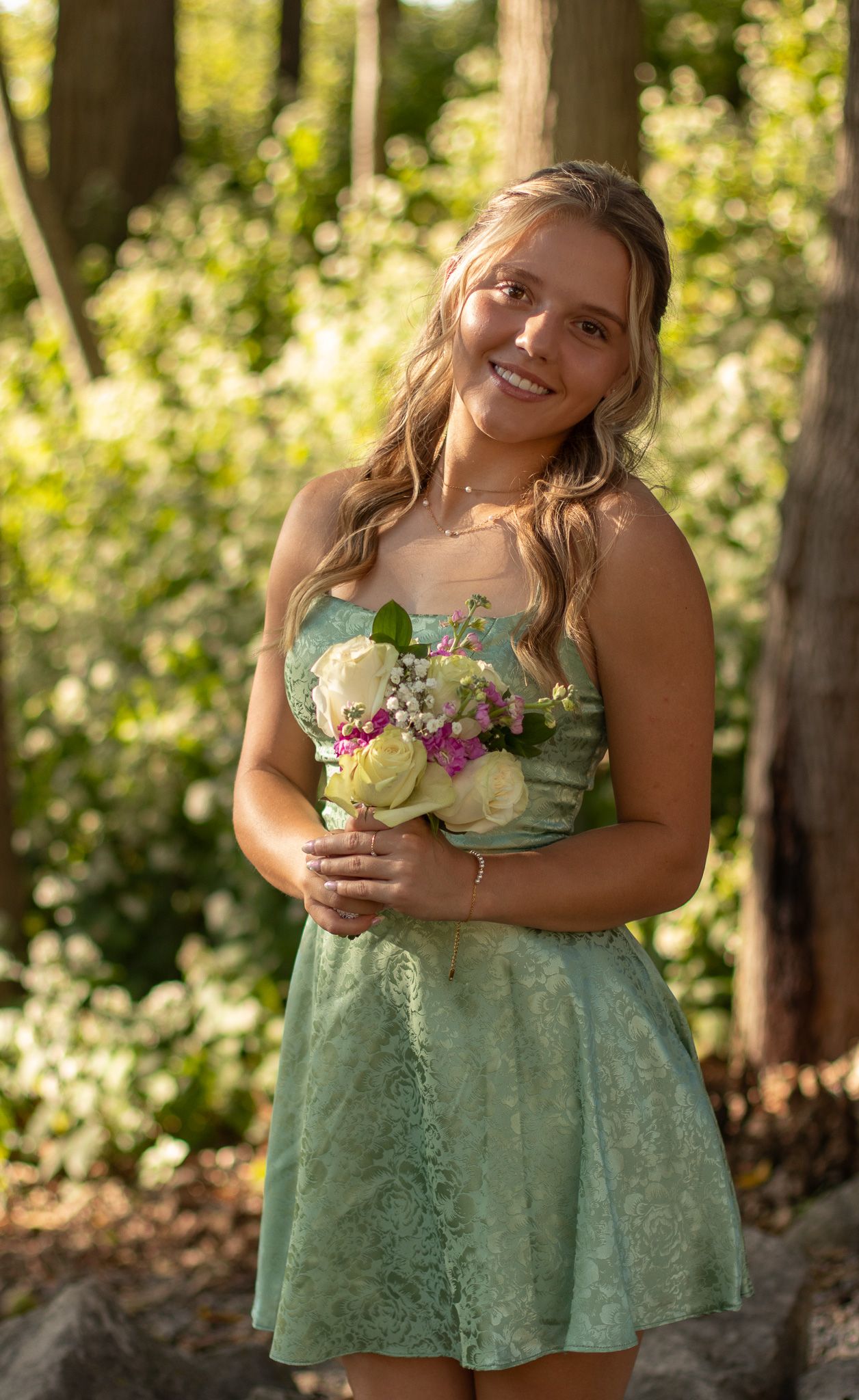 A person in a mint green lace dress holds a small bouquet while standing in dappled sunlight among trees.
